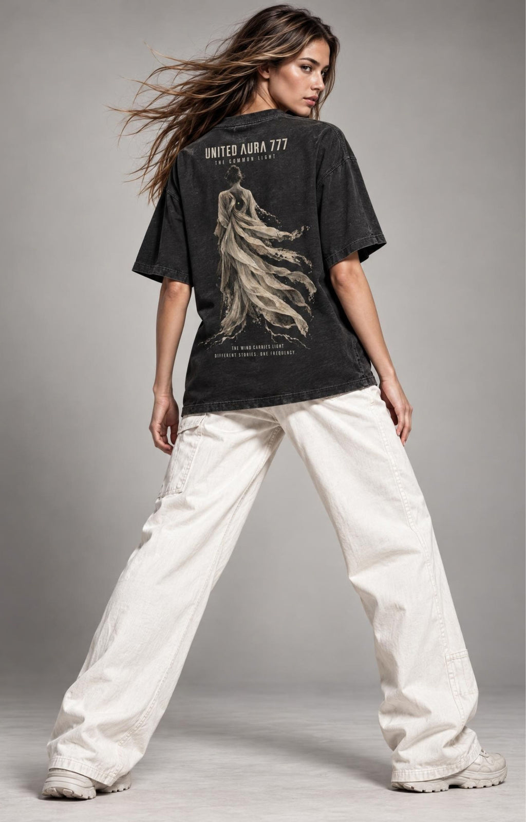 A woman modeling an oversized black graphic t-shirt with a beige print of a winged figure and the text 'UNITED ARBA 777' on the back, paired with wide-leg off-white pants, against a neutral gray studio background.