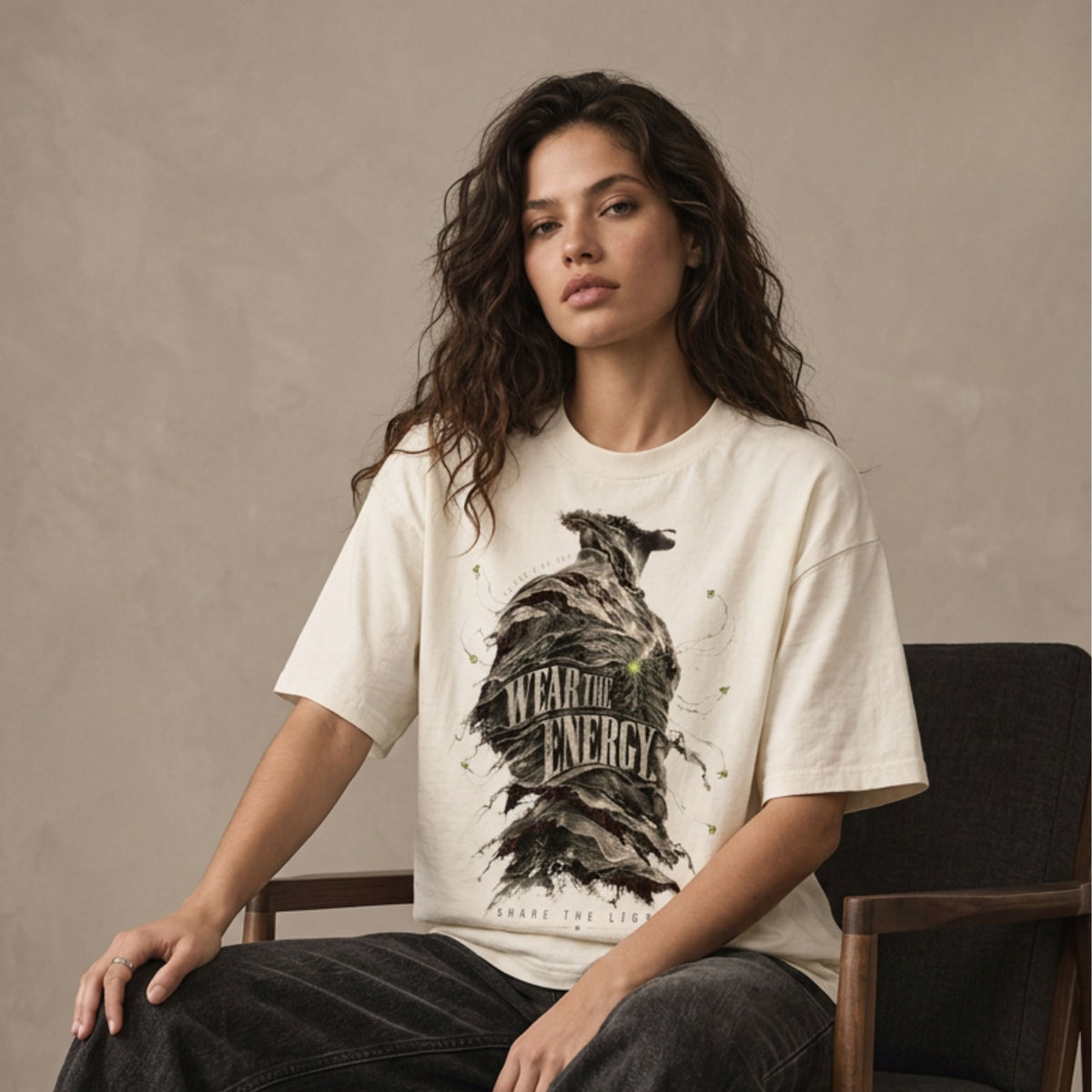 A woman sits on a wooden chair wearing a cream-colored graphic t-shirt with a black raven illustration and the text 'WEAR THE ENERGY', paired with dark gray wide-leg jeans, against a neutral textured studio backdrop.
