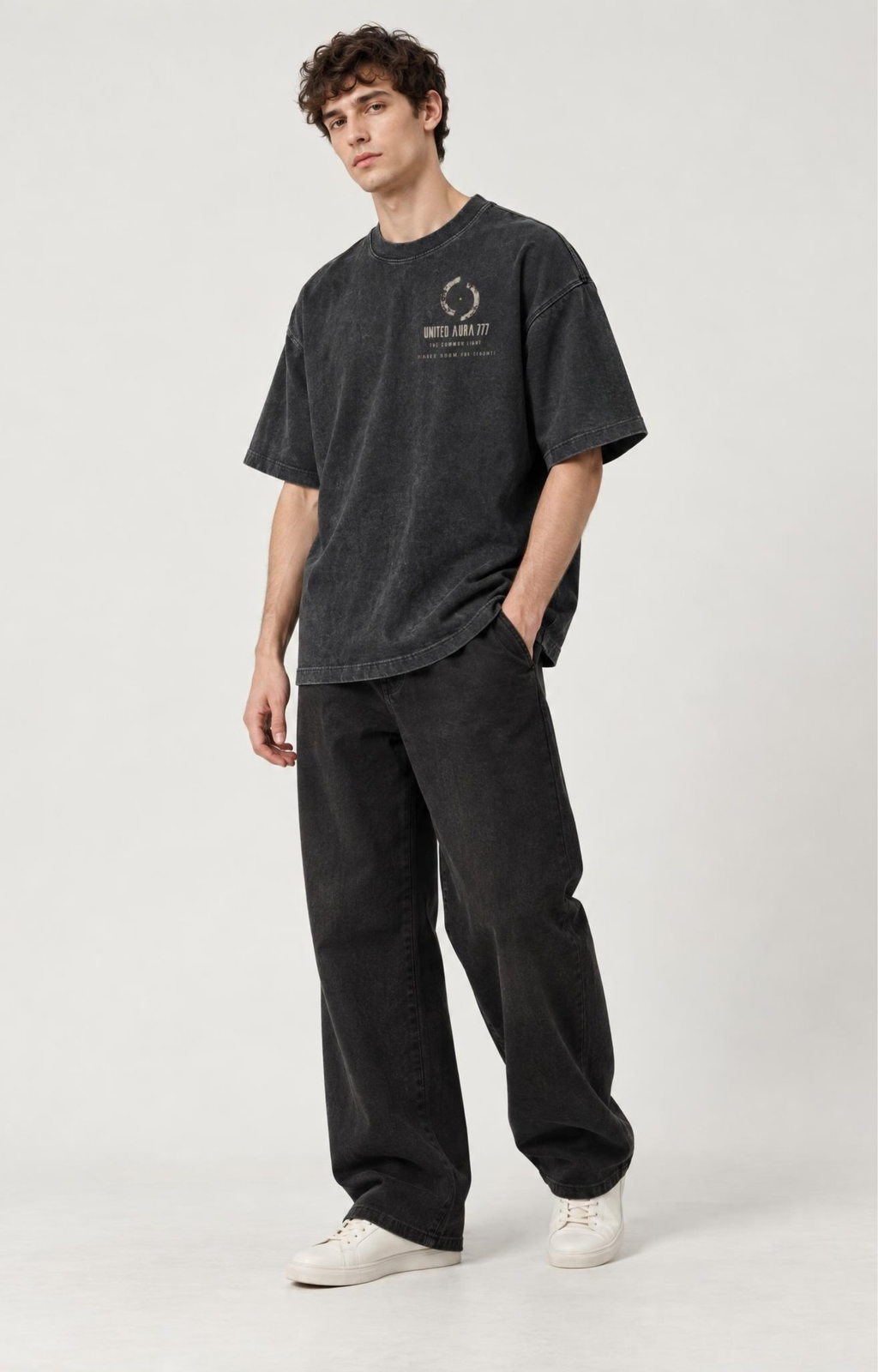 A man modeling an oversized dark gray washed t-shirt with a small graphic on the left chest, paired with black wide-leg jeans and white sneakers, against a light gray studio background.