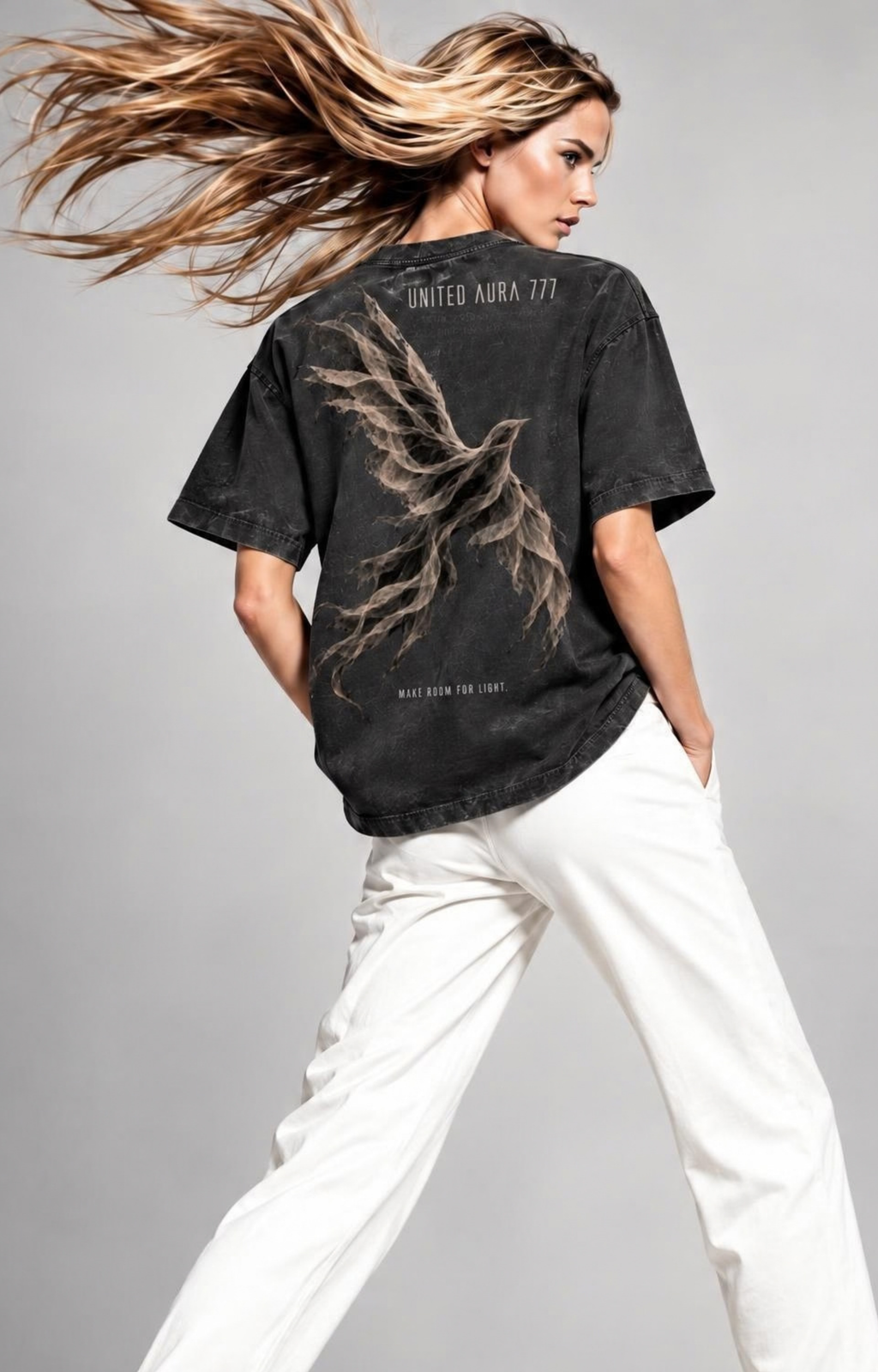 A woman wearing a faded black graphic t-shirt with a phoenix design and 'UNITED AURORA 777' text, paired with white pants, captured in a dynamic pose against a gray studio background.