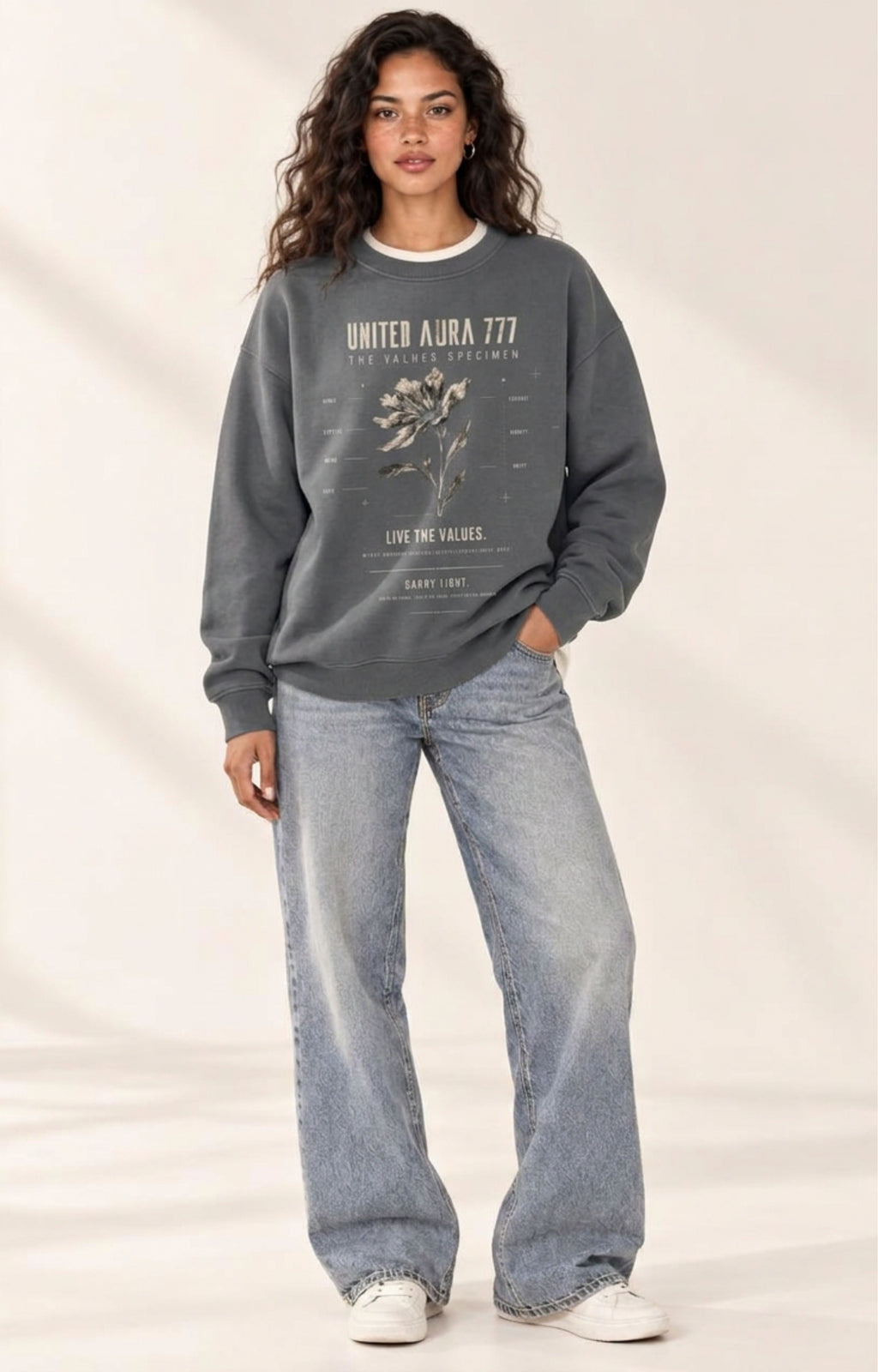 A woman modeling a dark gray crewneck sweatshirt with a vintage-style graphic print that reads 'UNITED AURA 777' and 'LIFE IS VALUABLE.' She is wearing light-wash wide-leg jeans and white sneakers against a warm beige studio background.