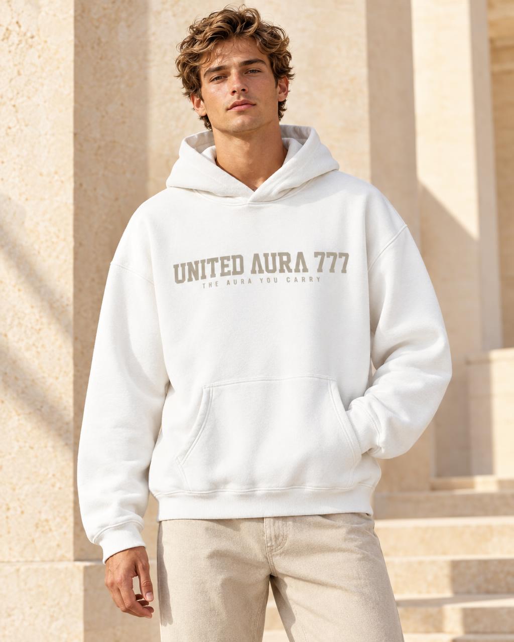 A man wearing a white pullover hoodie with 'UNITED AURA 777' printed in brown across the chest, paired with light beige pants, standing outdoors in front of a beige stone building.