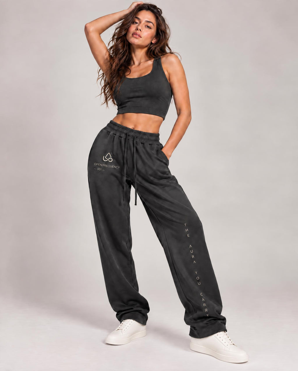 A woman modeling a dark gray two-piece activewear set consisting of a cropped tank top and matching jogger sweatpants, paired with white sneakers, against a neutral studio background.