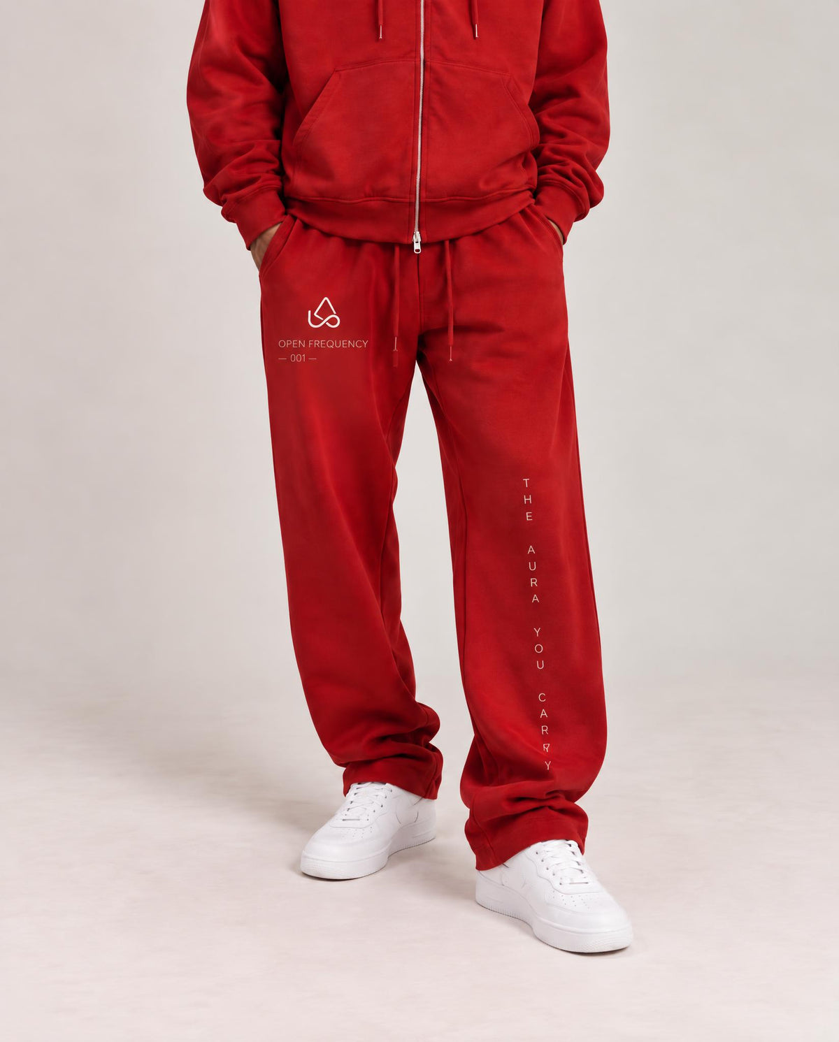 A person wearing a matching red zip-up hoodie and red sweatpants with white sneakers, standing against a light gray studio background. The sweatpants have white printed graphics on the left thigh and down the right leg.