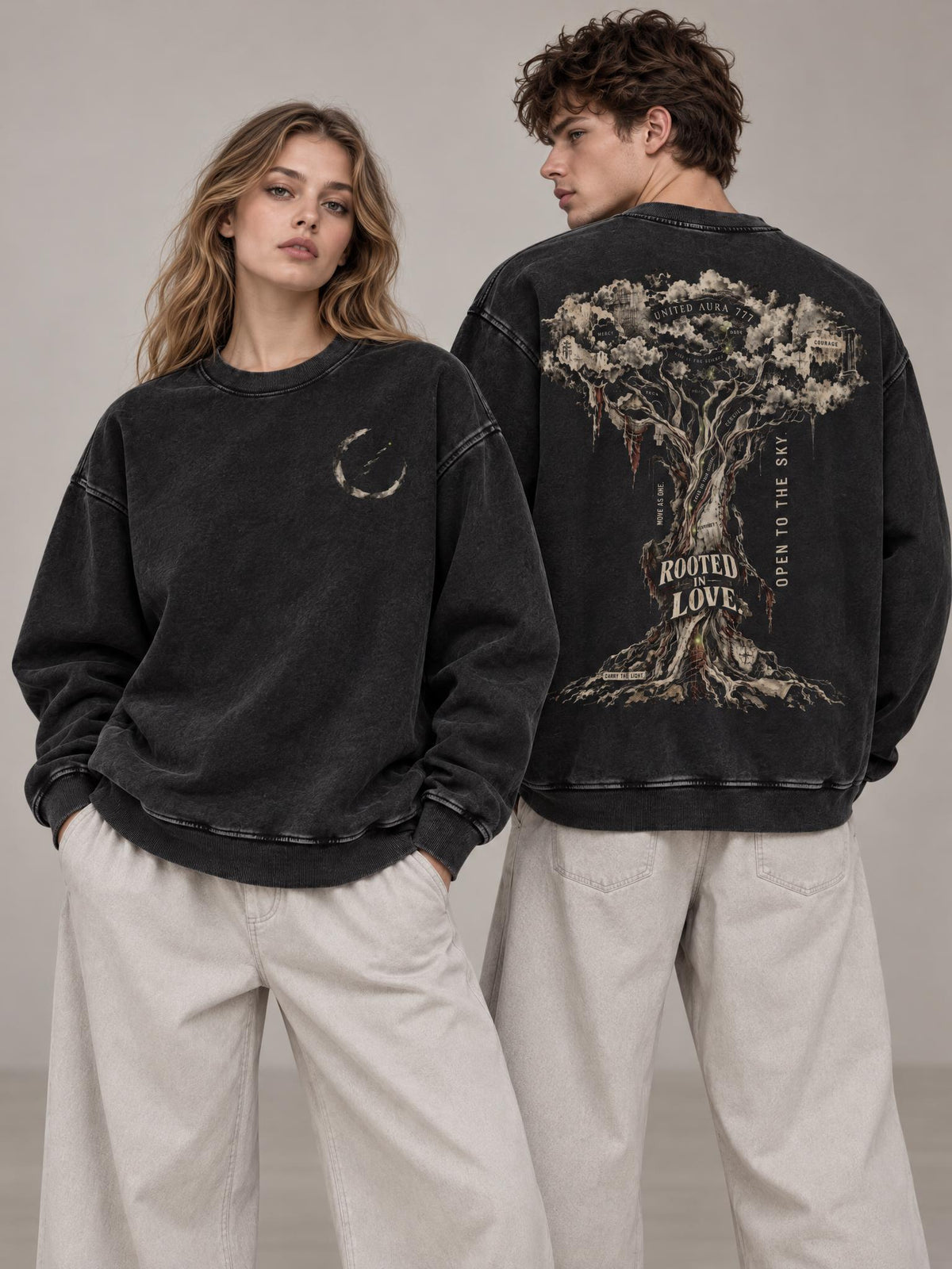 Two models wearing oversized black washed sweatshirts, one showing the front with a small moon graphic and the other showing the back with a large tree graphic and the words 'ROOTED LOVE' and 'GROW TO THE SKY', paired with light beige pants against a neutral studio background.
