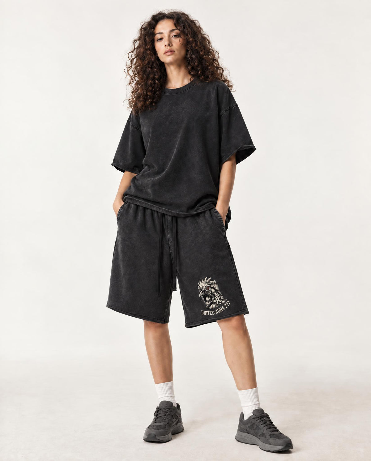 A model wearing a black mineral-wash oversized t-shirt and matching black mineral-wash shorts with a white graphic print on the left leg, styled with white socks and dark gray sneakers against a light off-white studio background.