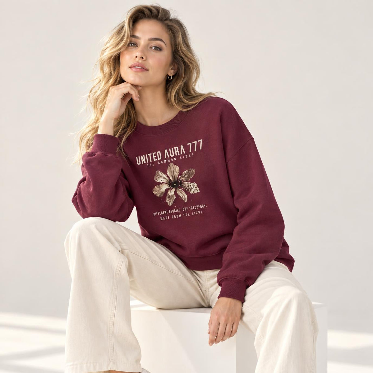 A woman sits on a white cube wearing a burgundy crewneck sweatshirt with a graphic print, cream wide-leg pants, and white sneakers in a bright studio setting.