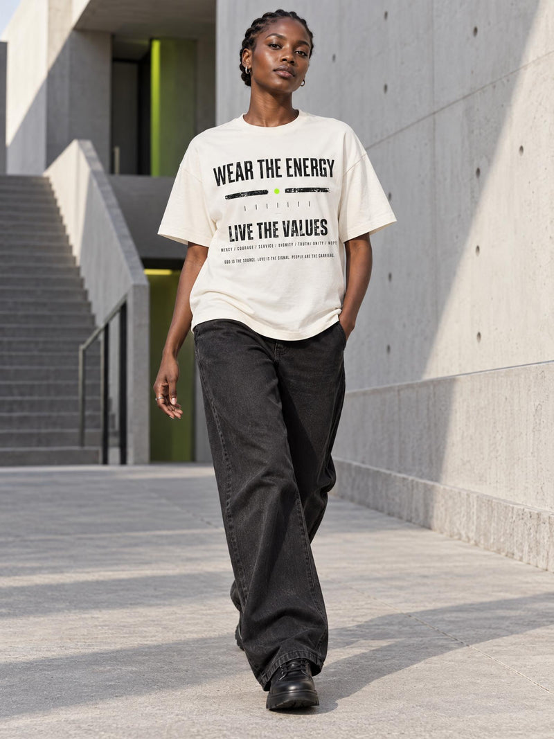 A model walking outdoors in a concrete corridor wearing an oversized cream-colored graphic t-shirt with black text that reads 'WEAR THE ENERGY' and 'LIVE THE VALUES', paired with loose black jeans and black shoes.