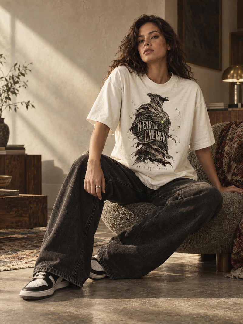 A woman sits on a textured armchair in a warm, sunlit room wearing an oversized white graphic T-shirt with a dark bird illustration and the words 'WEAR THE ENERGY,' paired with loose dark jeans and black-and-white sneakers.