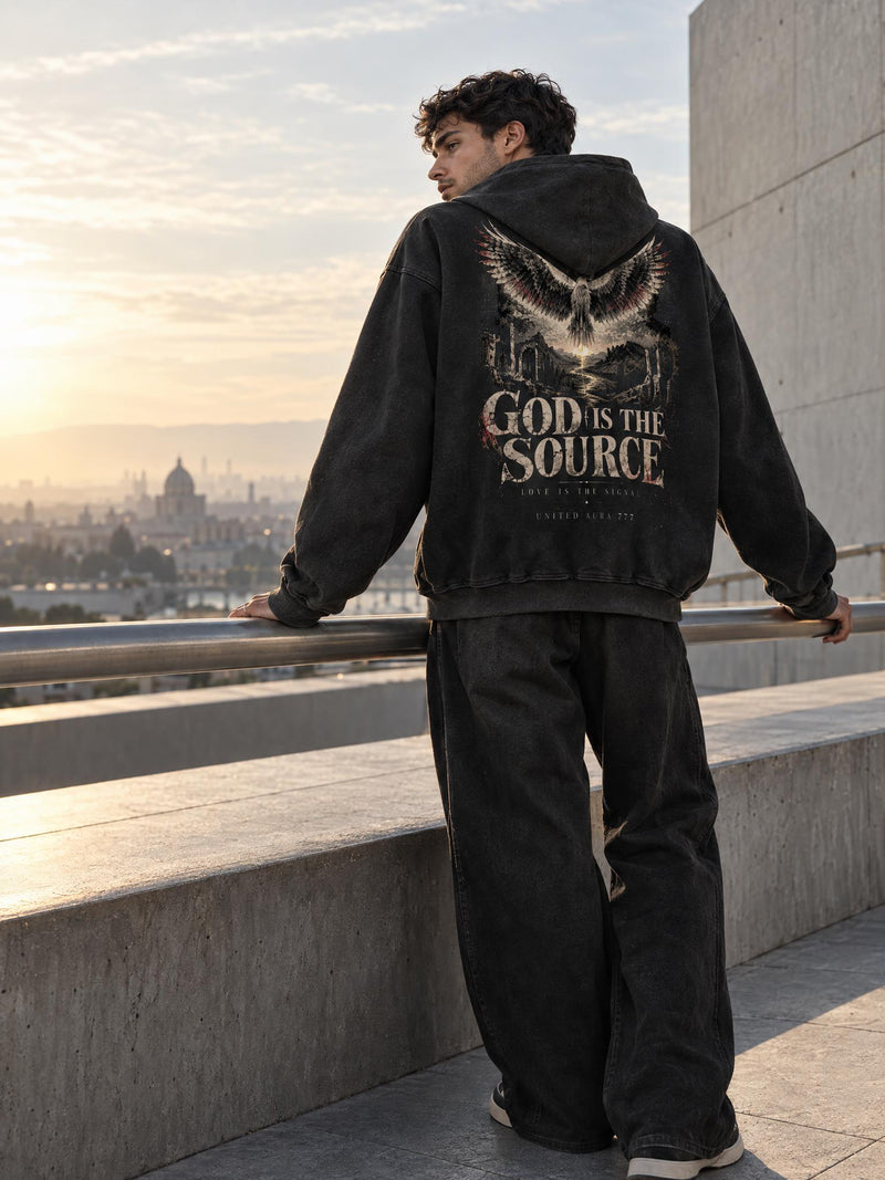 A man stands on an outdoor balcony at sunset wearing a faded black hoodie with a large graphic print on the back featuring an owl and the text 'GOD IS THE SOURCE'.