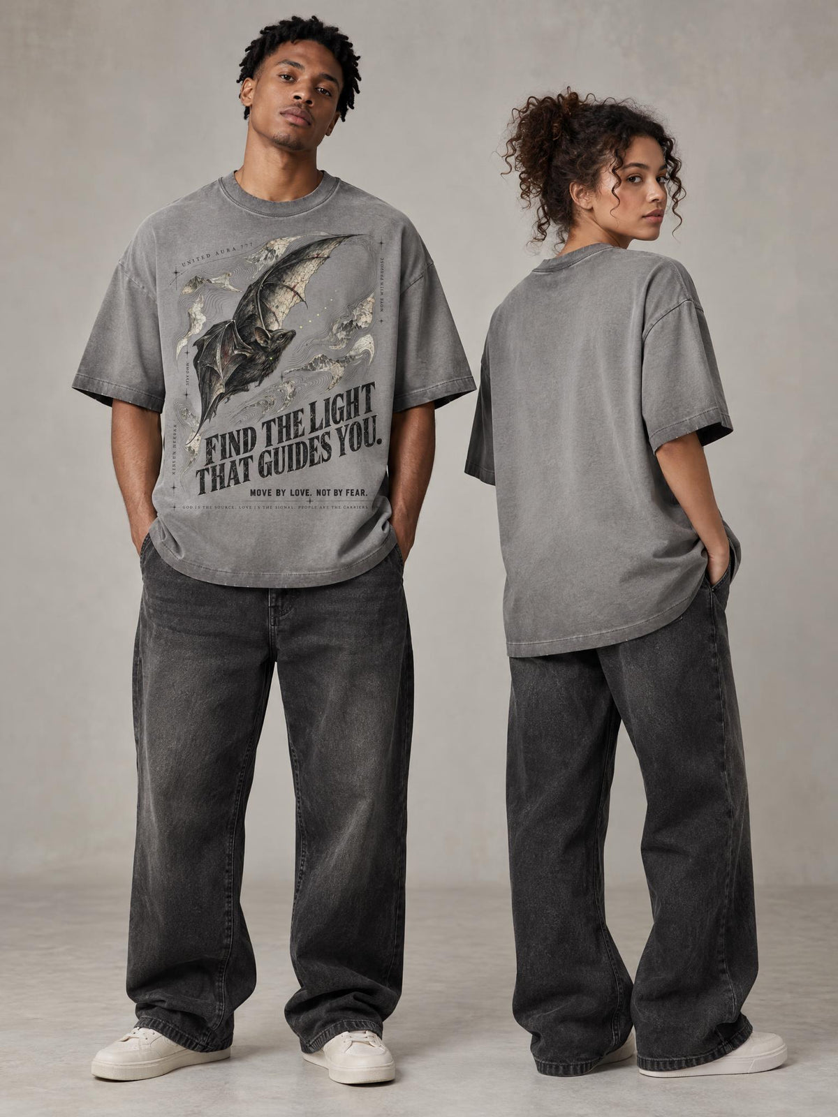 Two models wearing oversized gray graphic t-shirts and dark gray wide-leg jeans, standing against a neutral gray studio backdrop. The male model faces forward showing a bat graphic with the text 'FIND THE LIGHT THAT GUIDES YOU' and 'MADE BY LUXE, WORN BY ALL'.