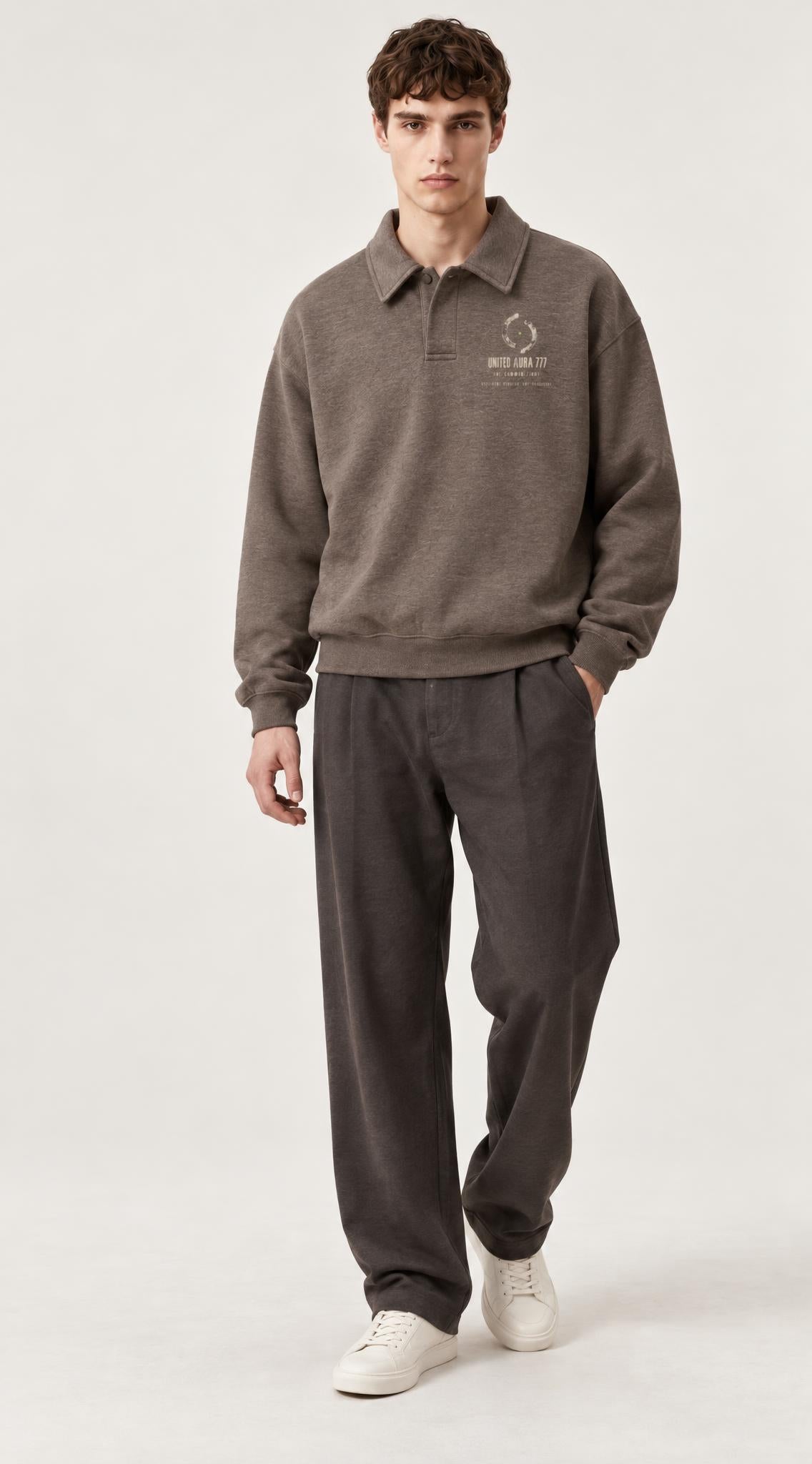 A male model wearing a brown long-sleeve polo sweatshirt with a small white graphic on the left chest, paired with dark gray pants and white sneakers, standing against a light gray studio background.