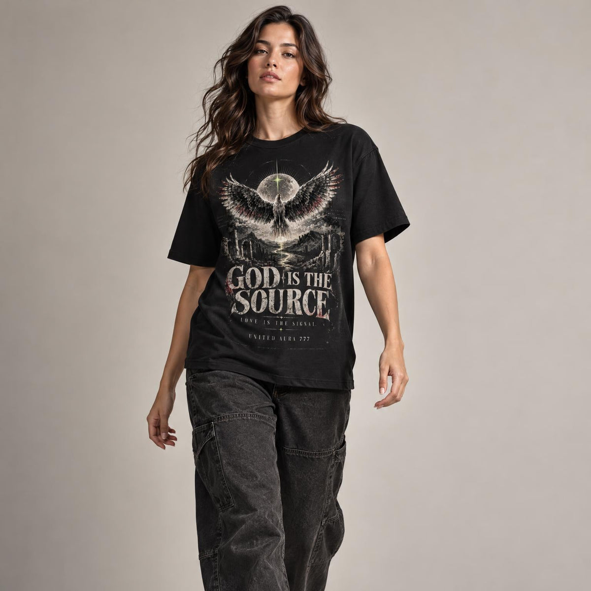 A woman modeling a black graphic t-shirt with a white and gray winged skull design and the text 'GOD IS THE SOURCE'. She is wearing loose black jeans and white sneakers against a neutral gray studio background.