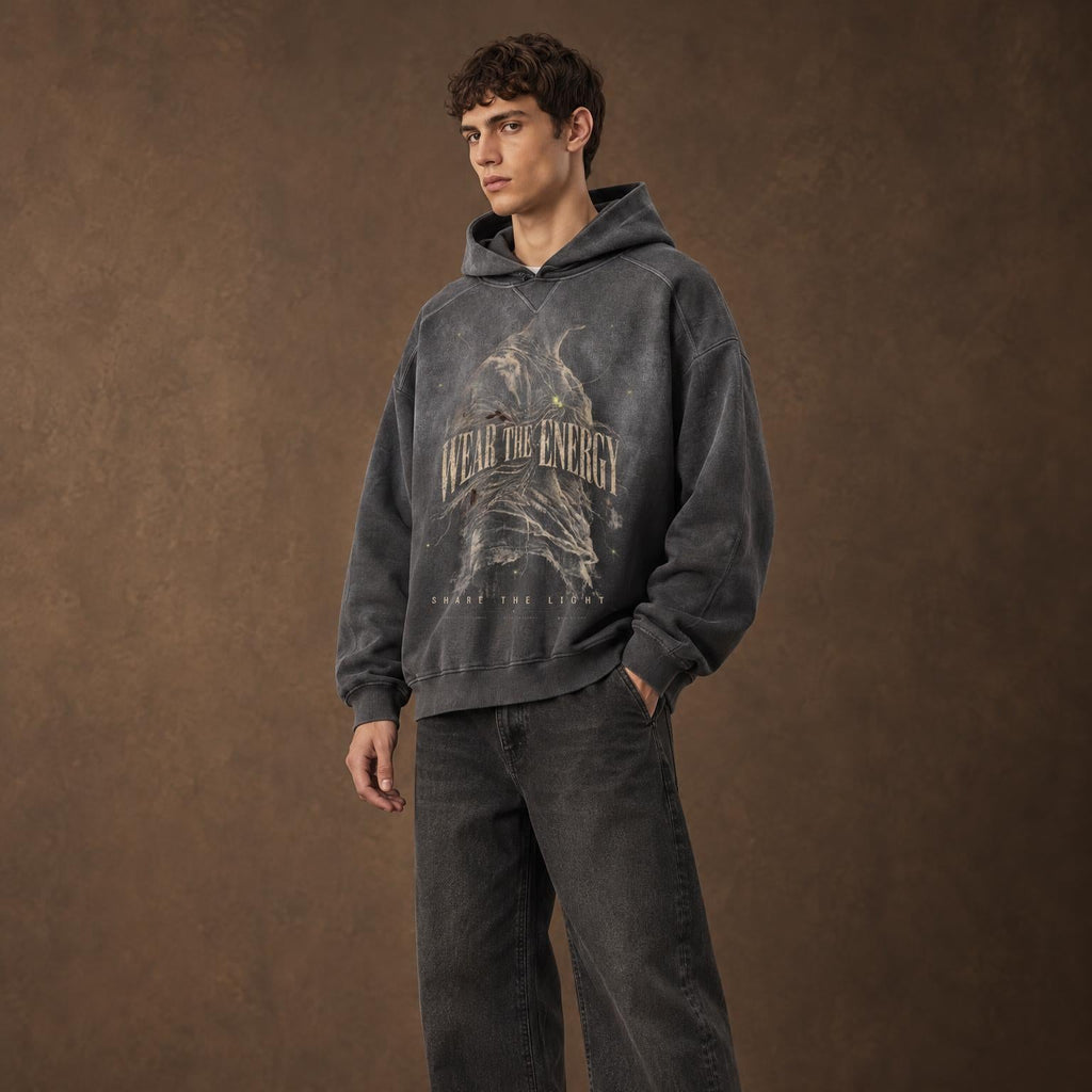 A man wearing a dark gray washed graphic hoodie with 'WEAR THE ENERGY' text and an eagle design, paired with dark gray jeans and black shoes, standing against a textured brown studio backdrop.