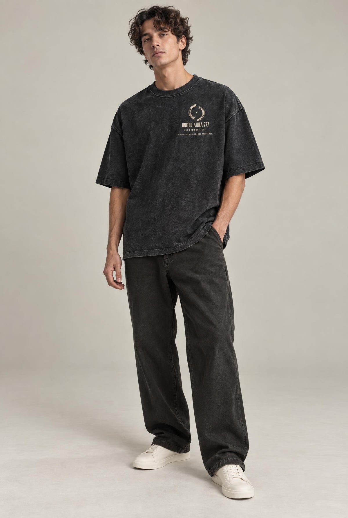 A man modeling an oversized black washed graphic t-shirt with a circular logo on the chest, paired with dark charcoal pants and white sneakers, against a neutral studio backdrop.