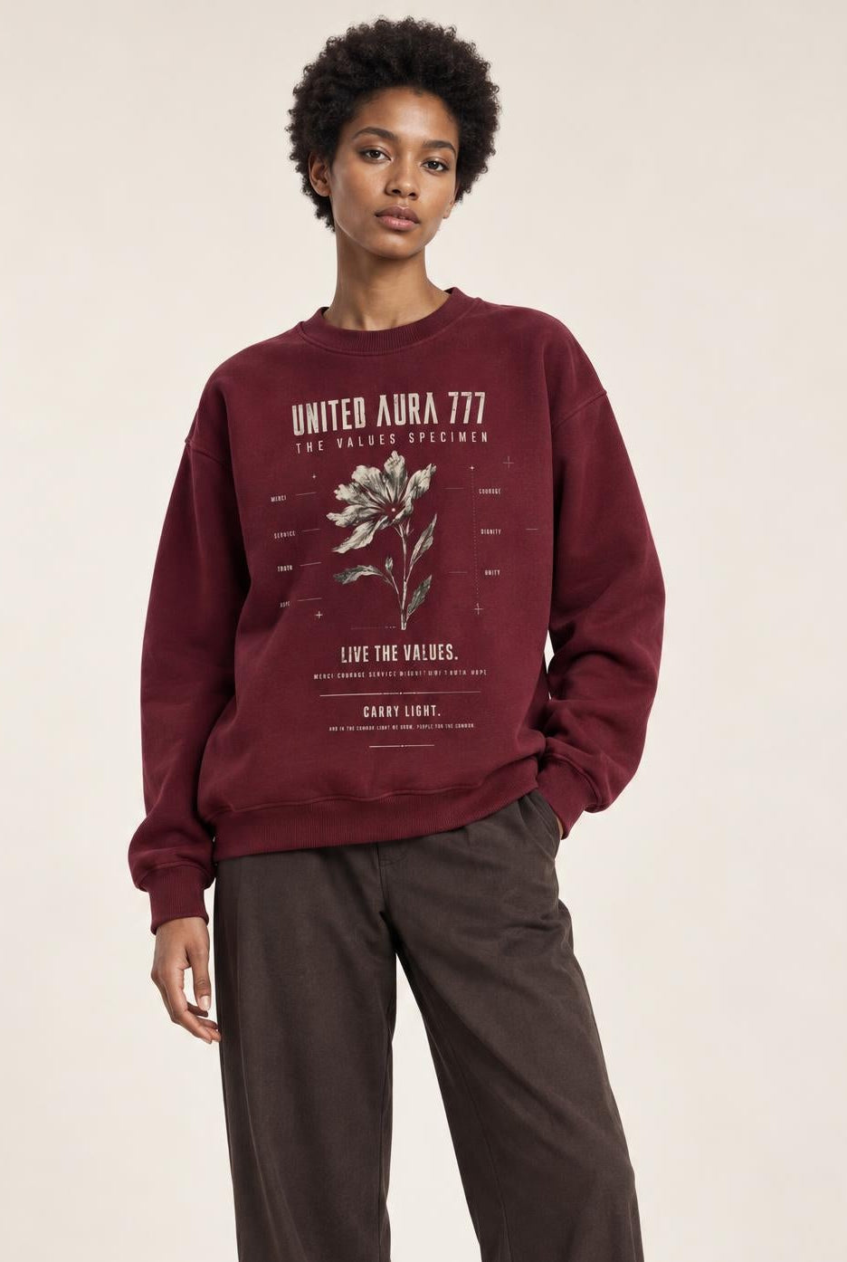 A model stands against a plain light beige studio background wearing a burgundy crewneck sweatshirt with a vintage graphic print, dark brown wide-leg trousers, and white sneakers.