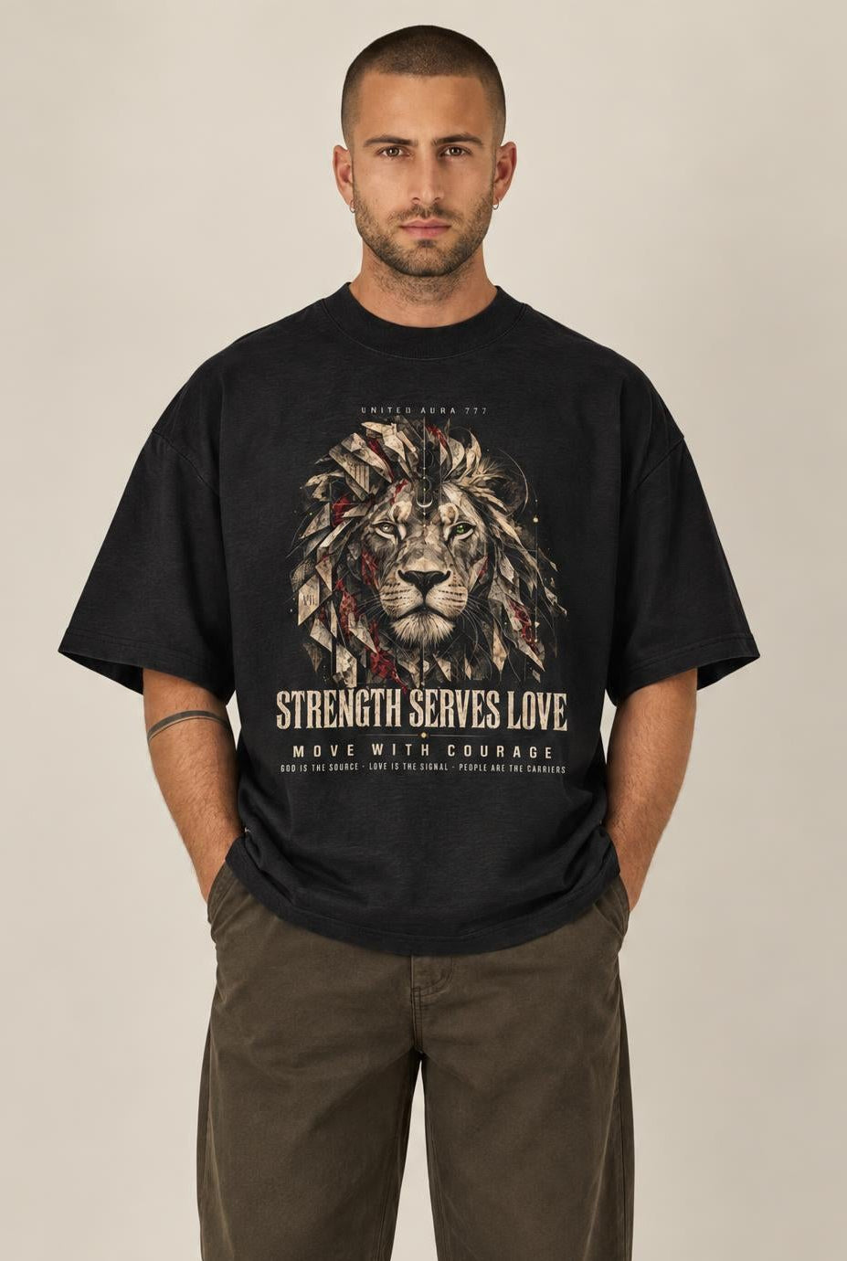 A man modeling a black graphic t-shirt with a lion print and the text 'STRENGTH SERVES LOVE' and 'GIVE WITH GREATER REWARDS'. He is wearing dark brown pants and white sneakers against a neutral beige background.