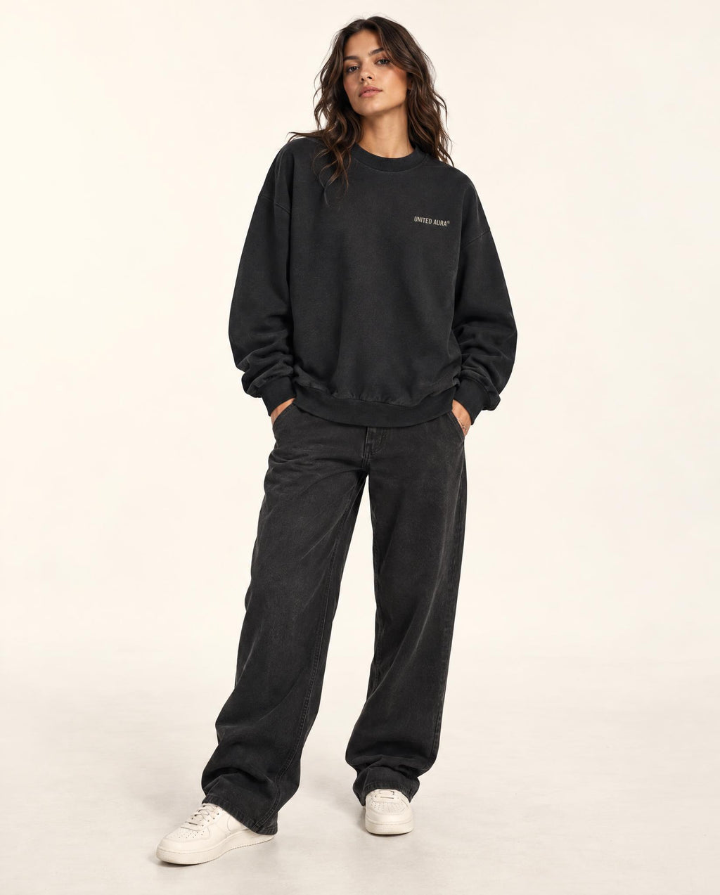 A woman modeling a black crewneck sweatshirt and matching black wide-leg sweatpants with white sneakers, standing against a plain off-white studio background.