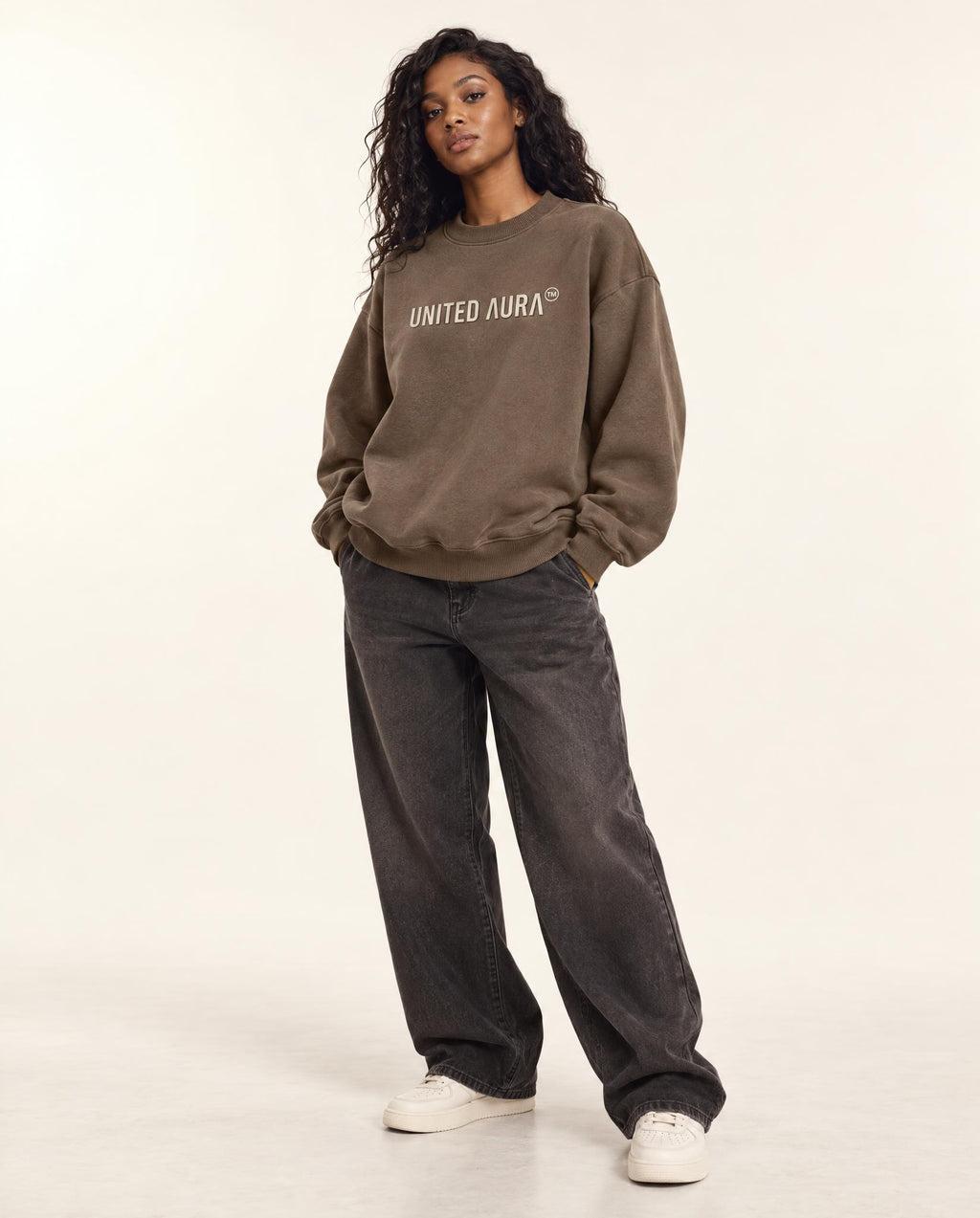 A model stands against a plain light background wearing a brown crewneck sweatshirt with 'UNITED AURA' printed in white across the chest, paired with dark gray wide-leg jeans and white sneakers.