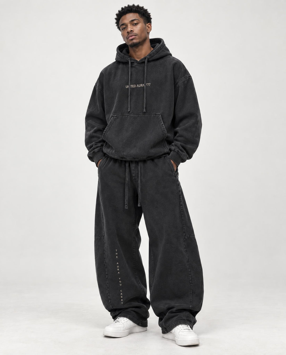 A man modeling a matching washed black hoodie and baggy black sweatpants with white sneakers, standing against a light gray studio background.