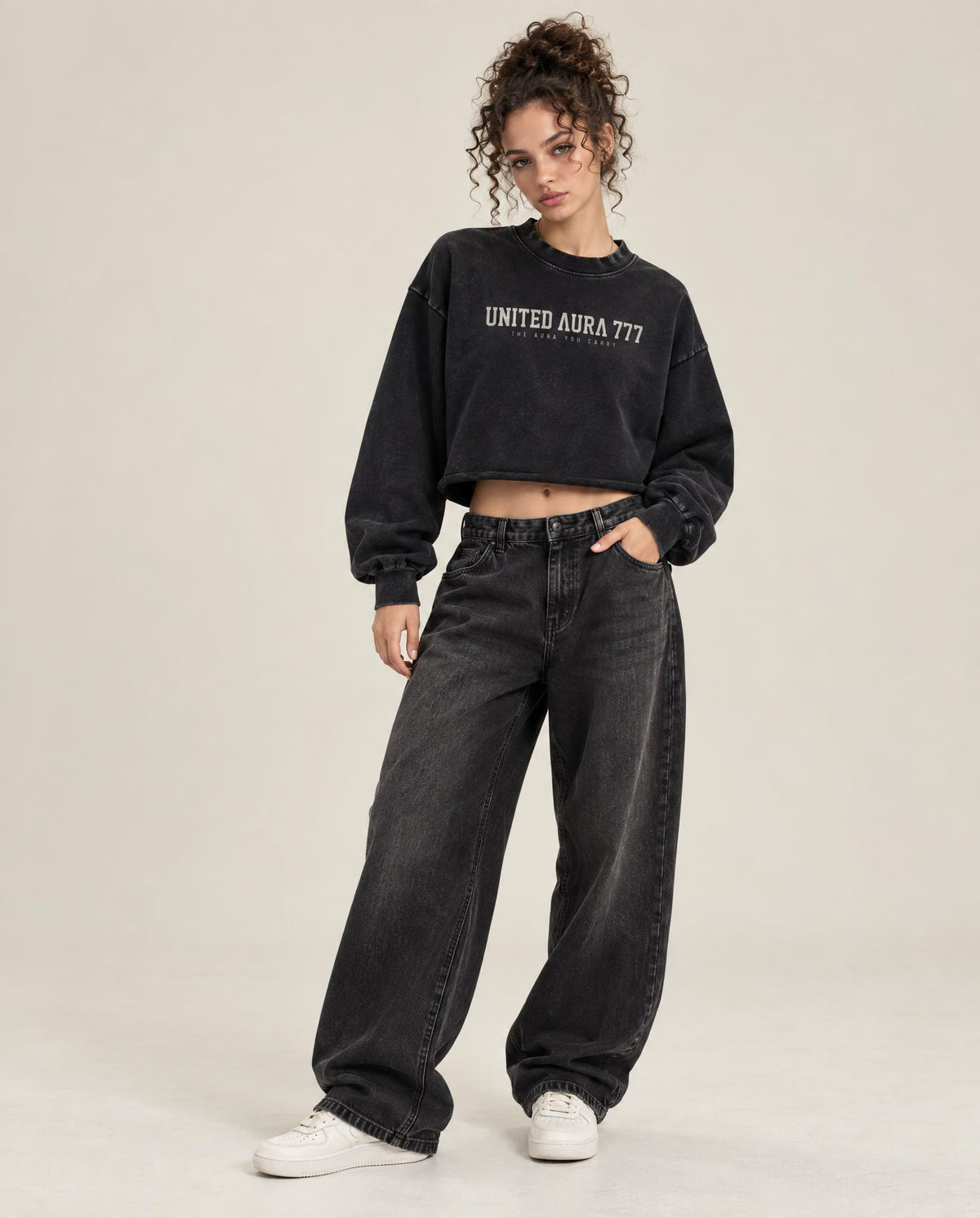 A model wearing a black cropped sweatshirt with 'UNITED AURA 777' printed in white on the chest, paired with wide-leg faded black jeans and white sneakers, against a light beige studio background.