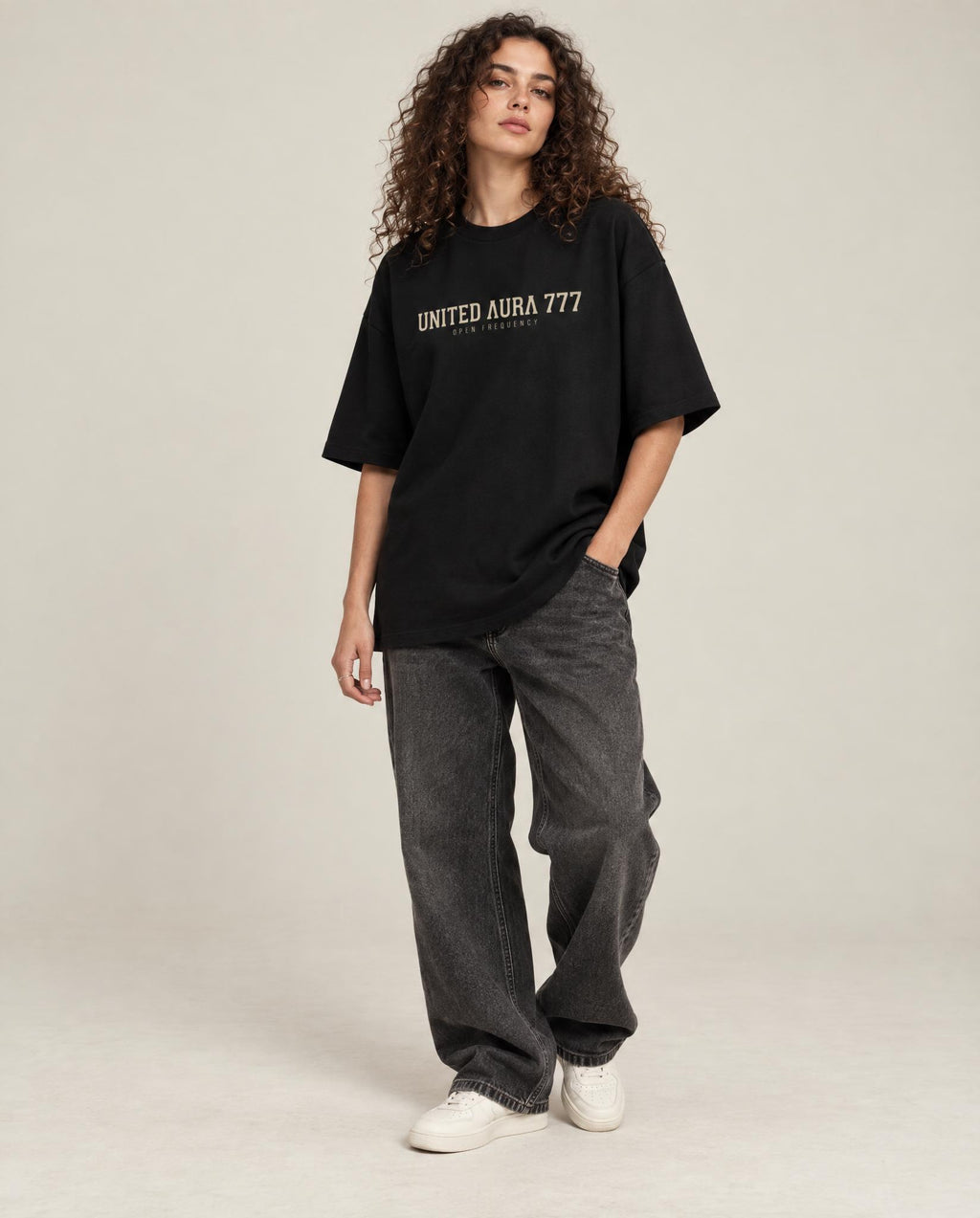 A model with curly brown hair stands against a light beige studio background wearing an oversized black t-shirt with 'UNITED AURA 777' printed in beige across the chest, paired with dark gray baggy jeans and white sneakers.