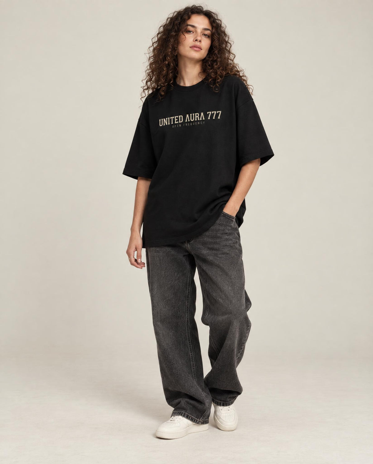A model with curly brown hair stands against a light beige studio background wearing an oversized black t-shirt with 'UNITED AURA 777' printed in beige across the chest, paired with dark gray baggy jeans and white sneakers.