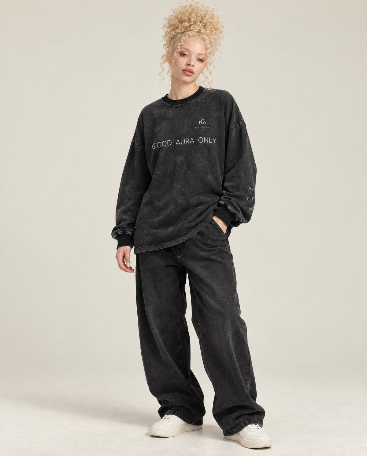 A model with curly blonde hair wearing a washed black long-sleeve sweatshirt with 'GOOD AURA ONLY' text and a small logo on the chest, paired with matching black sweatpants and white sneakers, against a light beige studio background.