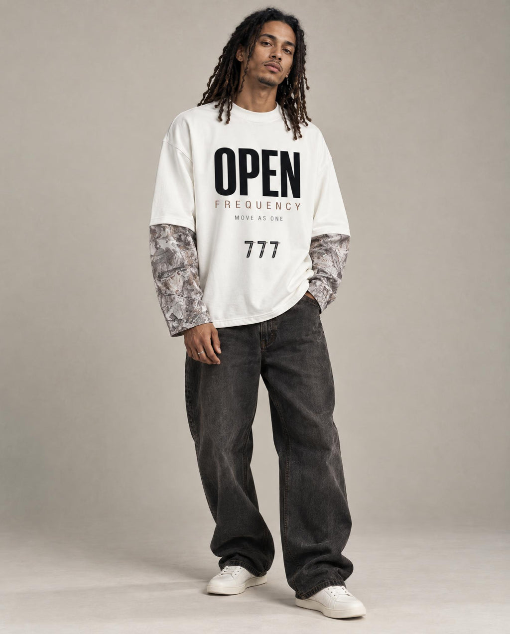 A model stands against a neutral studio backdrop wearing a white graphic T-shirt with black text reading 'OPEN FREQUENCY' and '777', layered over patterned long-sleeve undershirts, dark gray baggy jeans, and white sneakers.