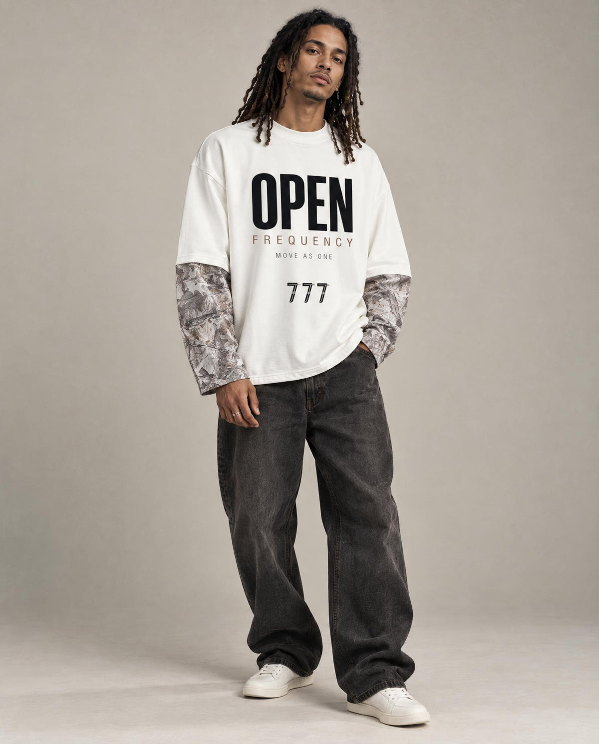 A model stands against a neutral studio backdrop wearing a white graphic T-shirt with black text reading 'OPEN FREQUENCY' and '777', layered over patterned long-sleeve undershirts, dark gray baggy jeans, and white sneakers.