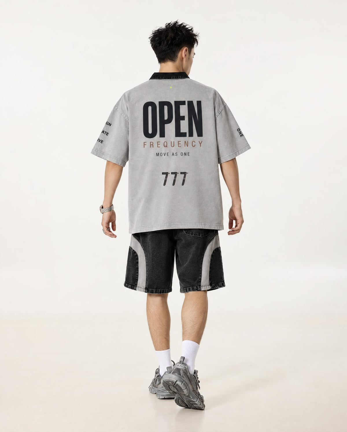 A man shown from behind wearing a light gray oversized t-shirt with 'OPEN FREQUENCY' printed in black on the back, paired with black and gray shorts, white socks, and gray sneakers, against a plain off-white studio background.