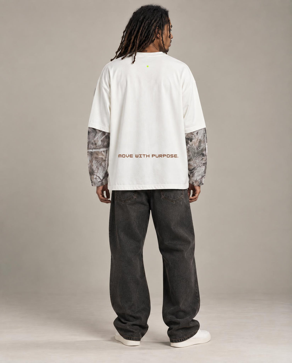 A model viewed from the back wearing a white long-sleeve t-shirt with patterned sleeves, dark gray baggy jeans, and white sneakers against a neutral studio backdrop.