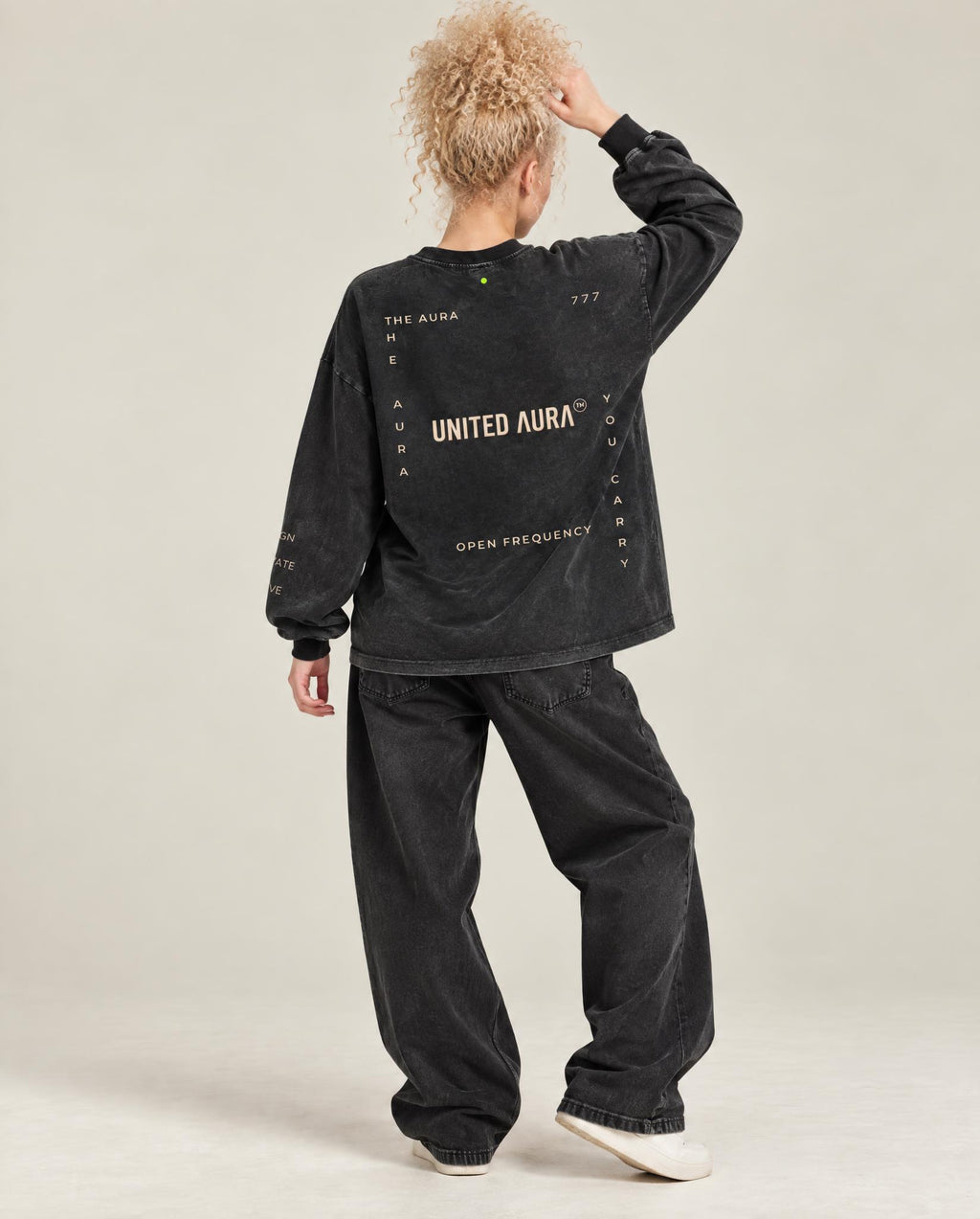 A model from behind wearing a washed black long-sleeve graphic sweatshirt and matching washed black baggy jeans, standing against a neutral light gray studio background.