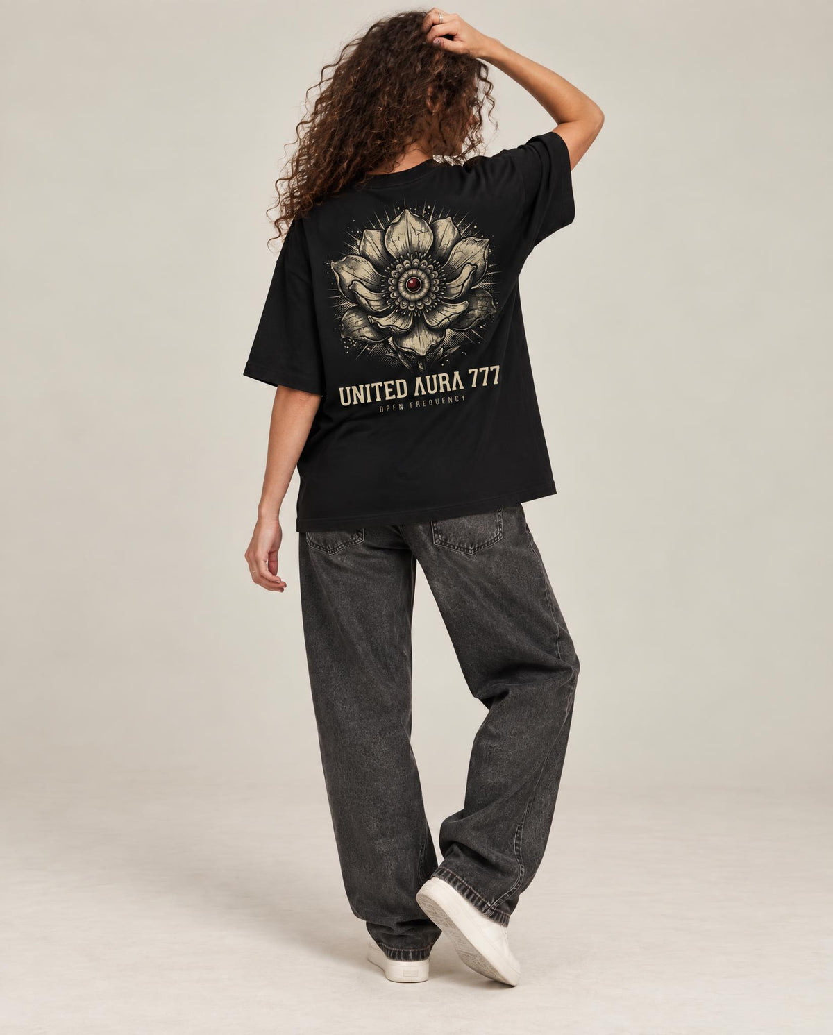 A model is shown from behind wearing a black oversized t-shirt with a large beige floral mandala graphic and the text 'UNITED AURA 777' on the back, paired with dark gray baggy jeans and white sneakers, against a light gray studio background.