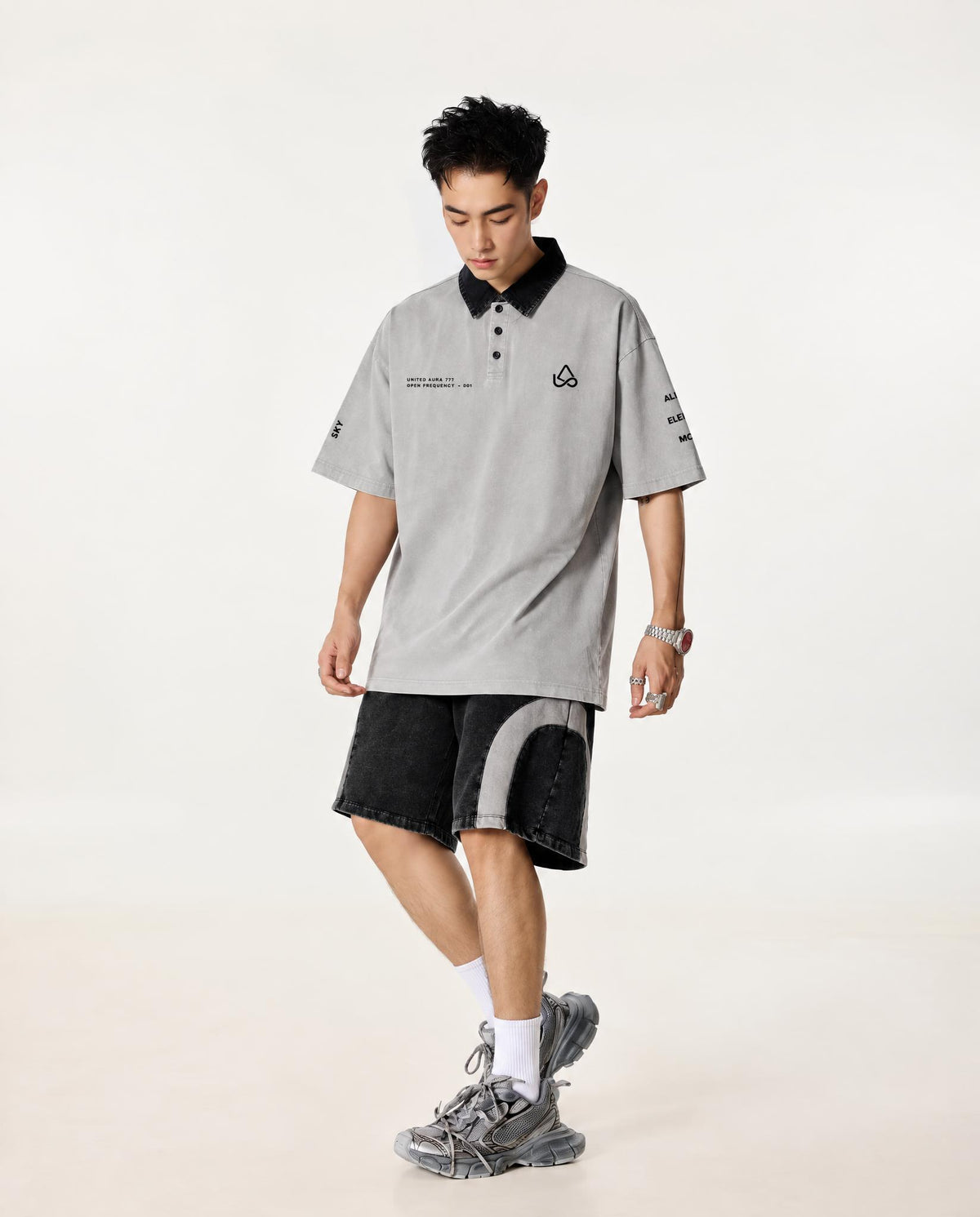 A man modeling a light gray oversized short-sleeve polo shirt with a black collar, paired with black and gray shorts, white socks, and gray sneakers, set against a plain off-white studio background.