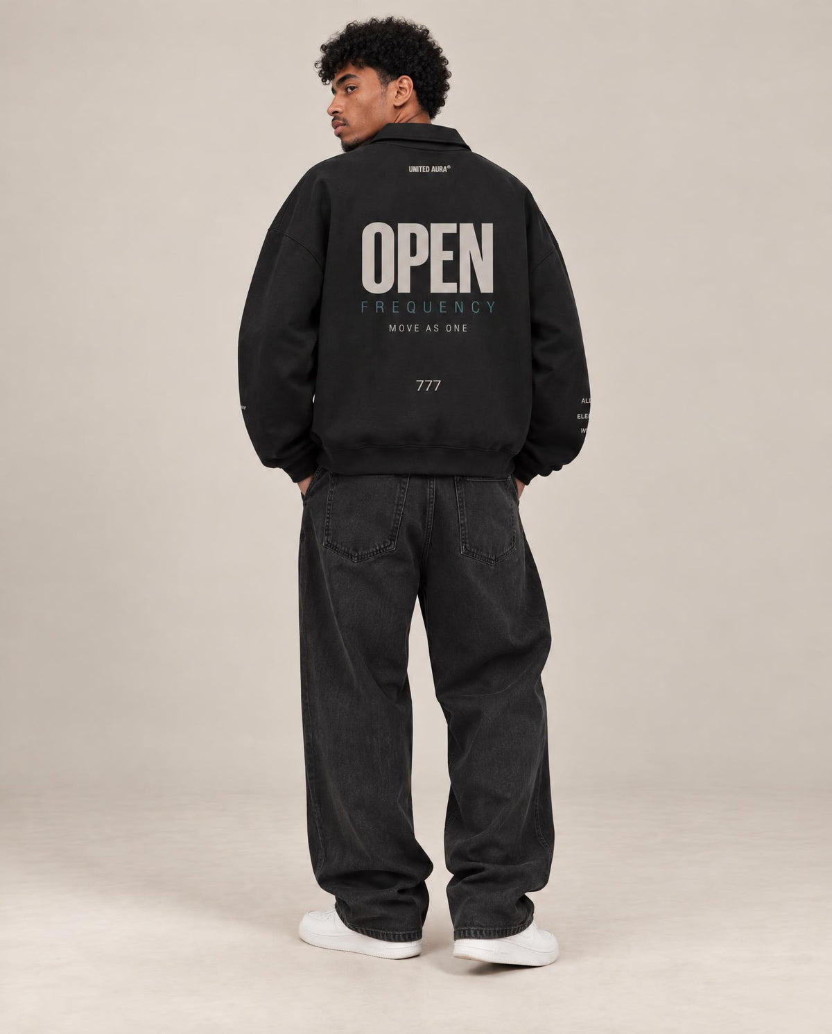 A man wearing a black oversized jacket with 'OPEN FREQUENCY' printed in white on the back, paired with dark jeans and white sneakers, photographed against a plain beige studio background.