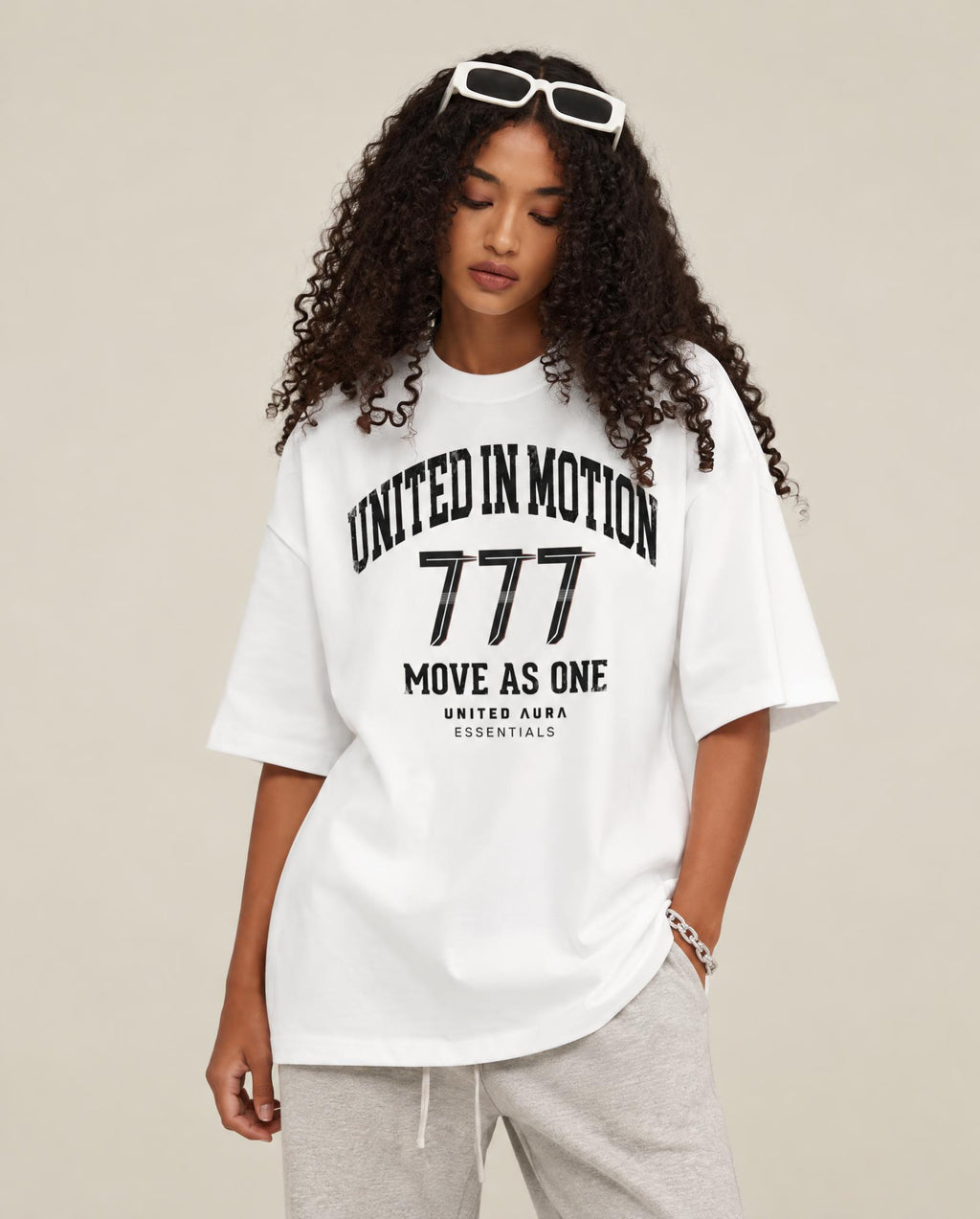 A woman wearing an oversized white graphic T-shirt with black text reading 'UNITED IN MOTION 777 MOVE AS ONE' and 'UNITED AURA ESSENTIALS', paired with light gray sweatpants against a plain beige studio background.