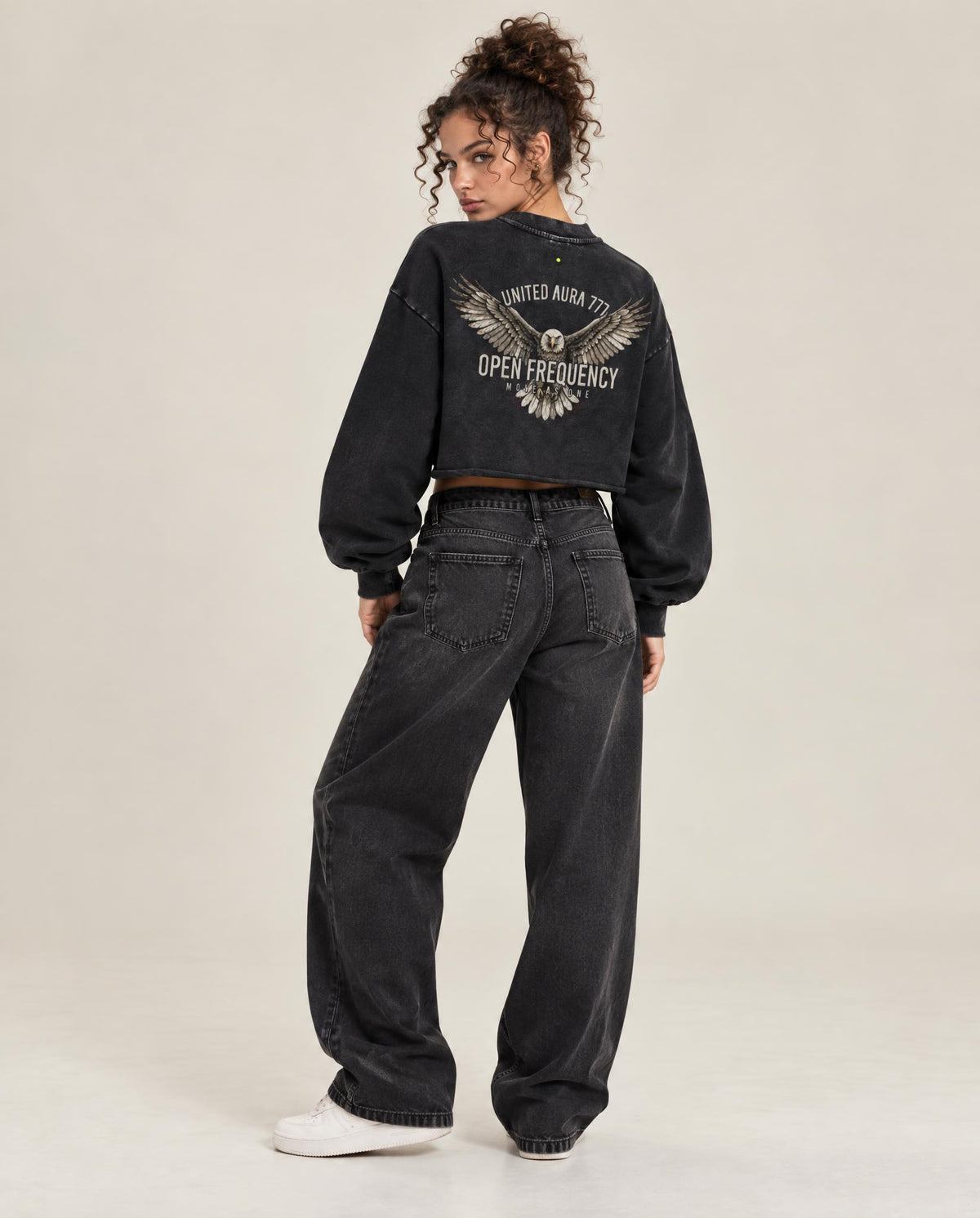 A model shown from the back wearing a black cropped sweatshirt with a large graphic print on the back, paired with dark gray wide-leg jeans and white sneakers, against a plain light beige studio background.