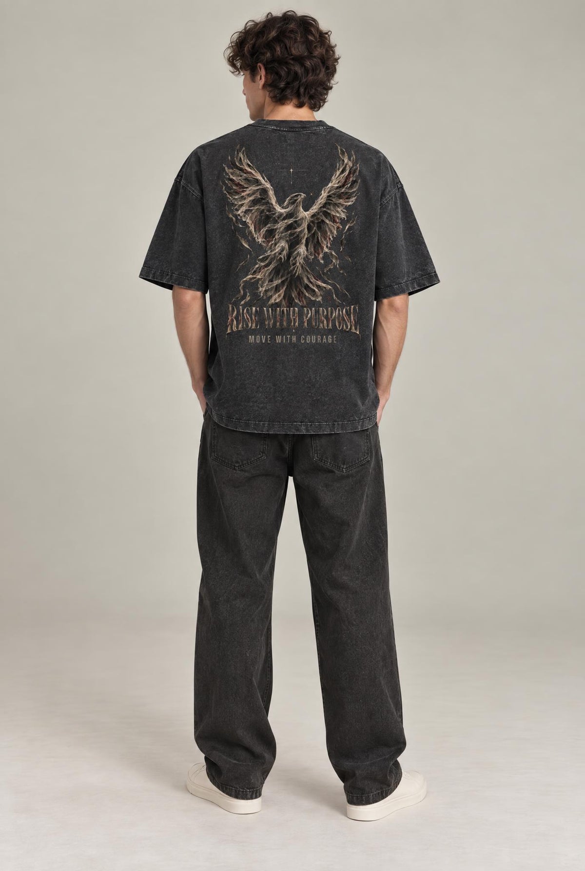 A man stands with his back to the camera wearing a faded black graphic t-shirt with a large eagle design and the text 'RISE WITH PURPOSE' on the back, paired with dark gray jeans and white sneakers, against a neutral studio backdrop.