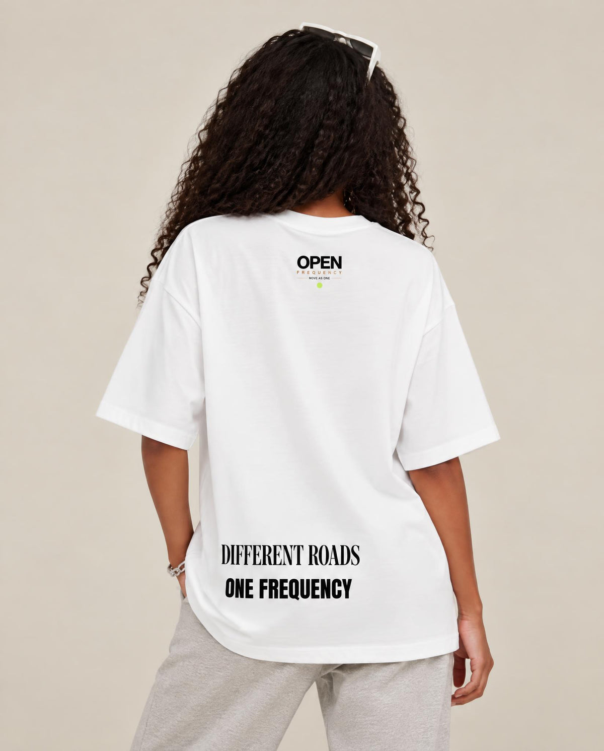 A woman shown from the back wearing an oversized white t-shirt with black text on the back reading 'OPEN' and 'DIFFERENTROADS ONE FREQUENCY', paired with light gray sweatpants against a solid beige background.