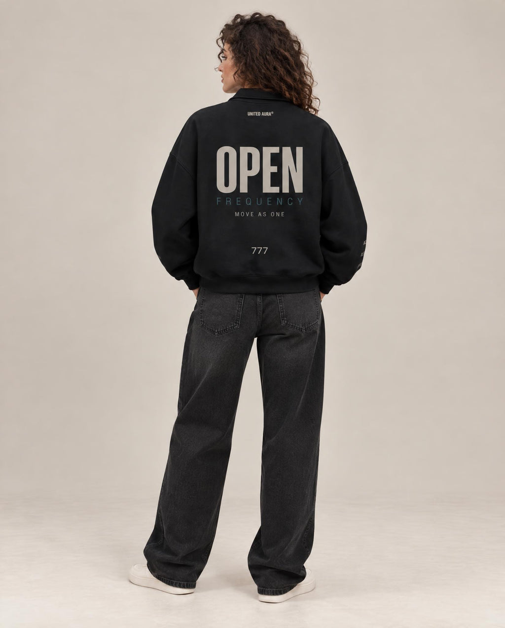 A model viewed from behind wearing a black bomber jacket with 'OPEN' printed in white on the back, paired with dark gray jeans and white sneakers, against a neutral studio background.