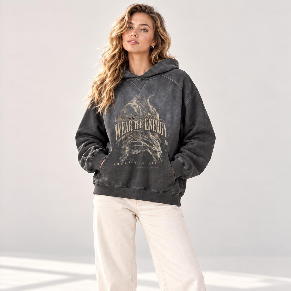 A woman modeling a dark gray washed graphic hoodie with 'HELL OR BURN' text, paired with cream wide-leg pants and white sneakers, standing in a bright studio setting.