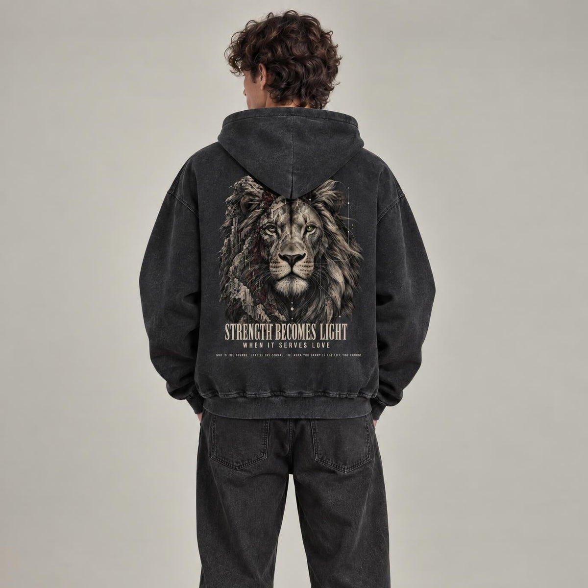 A model viewed from behind wearing a washed black hoodie with a large graphic of a lion on the back and the text 'STRENGTH BECOMES LIGHT' below it, paired with matching washed black sweatpants and white sneakers, against a neutral studio background.