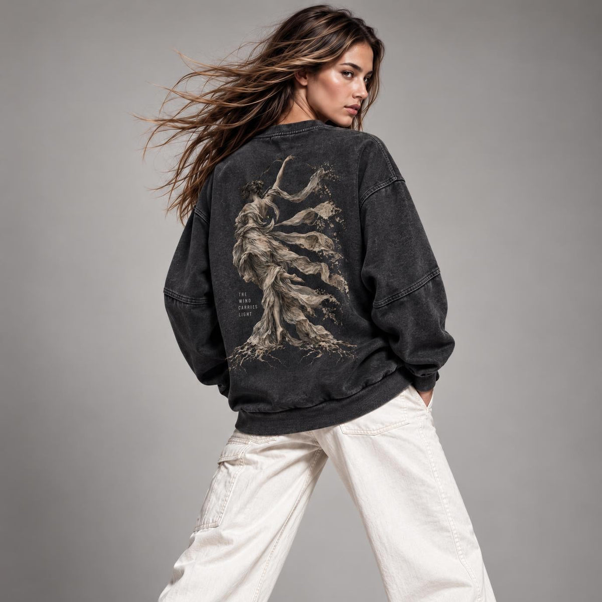 A woman modeling a washed black crewneck sweatshirt from the back, featuring a large graphic print of a winged figure on the back. She is wearing off-white wide-leg cargo pants and white sneakers against a neutral gray studio background.