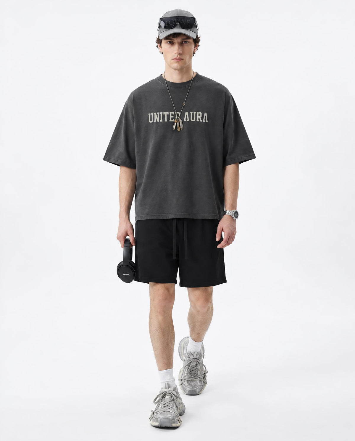 A male model walks forward in a studio setting wearing a dark charcoal gray oversized t-shirt with 'UNITED AURA' printed in white across the chest, paired with black athletic shorts, gray sneakers, and a gray cap.