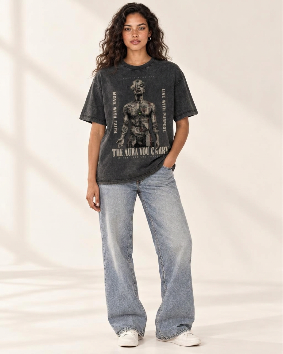 A woman modeling a dark charcoal gray graphic t-shirt with a black print of a classical statue and text, paired with light-wash wide-leg jeans and white sneakers, set against a warm beige background with soft shadows.