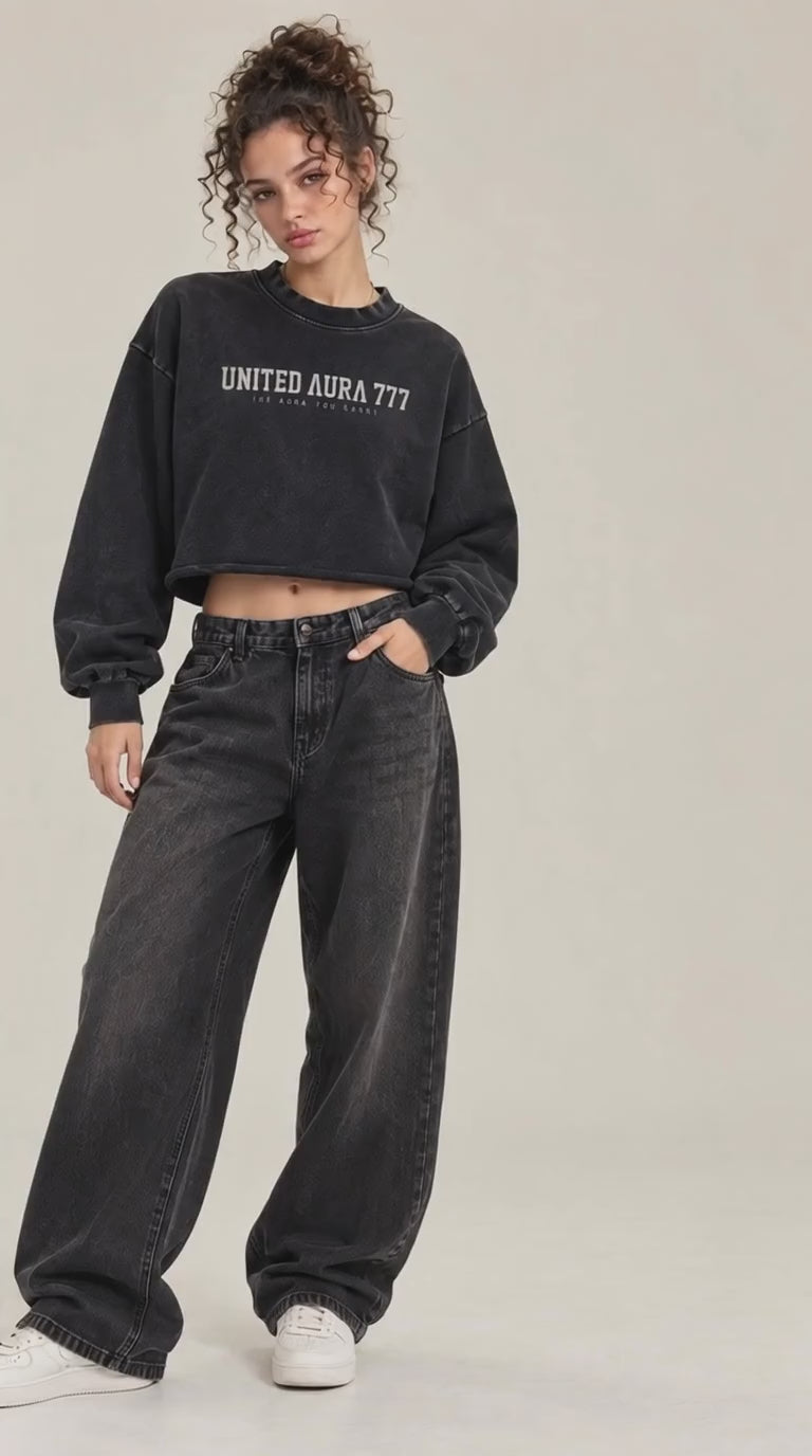 a video of A model shown from the back wearing a black cropped sweatshirt with a large graphic print on the back, paired with dark gray wide-leg jeans and white sneakers, against a plain light beige studio background.