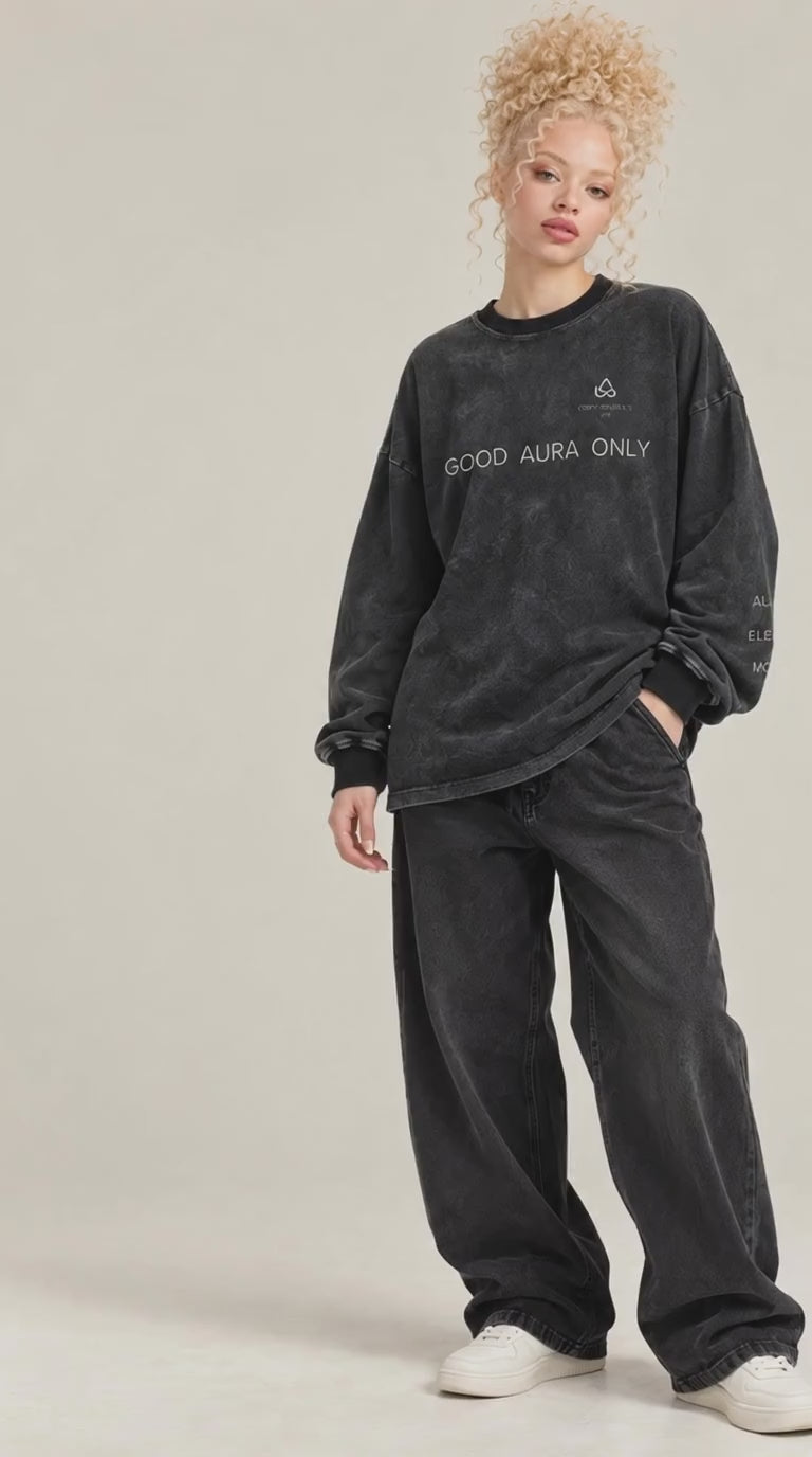 video of A model with curly blonde hair wearing a washed black long-sleeve sweatshirt with 'GOOD AURA ONLY' text and a small logo on the chest, paired with matching black sweatpants and white sneakers, against a light beige studio background.