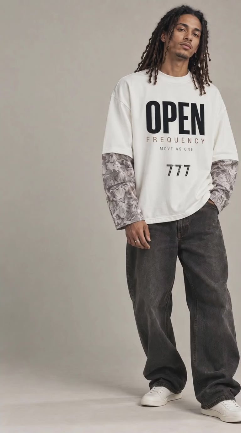 a video of A model stands against a neutral studio backdrop wearing a white graphic T-shirt with black text reading 'OPEN FREQUENCY' and '777', layered over patterned long-sleeve undershirts, dark gray baggy jeans, and white sneakers.