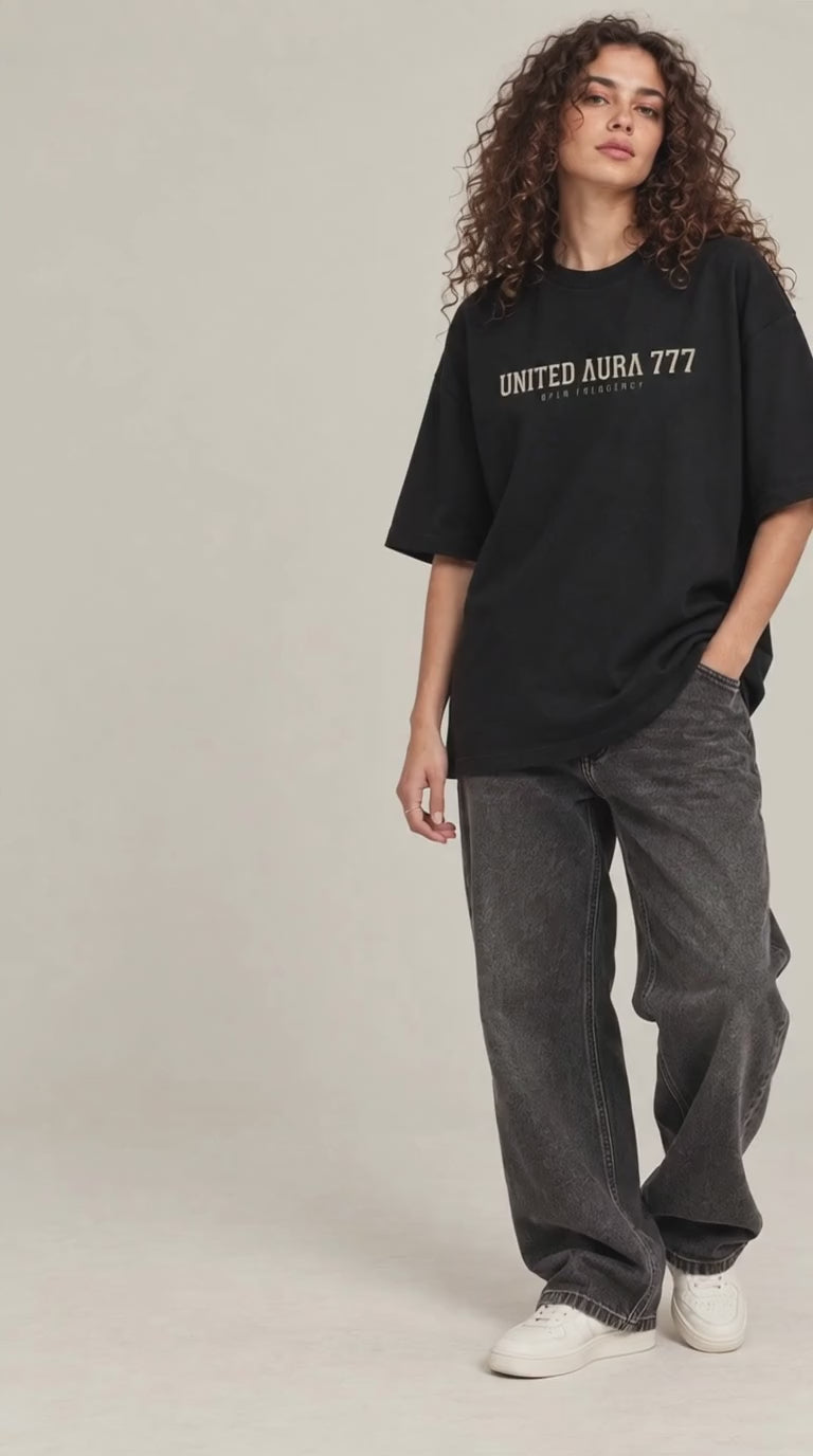 video of A model with curly brown hair stands against a light beige studio background wearing an oversized black t-shirt with 'UNITED AURA 777' printed in beige across the chest, paired with dark gray baggy jeans and white sneakers.